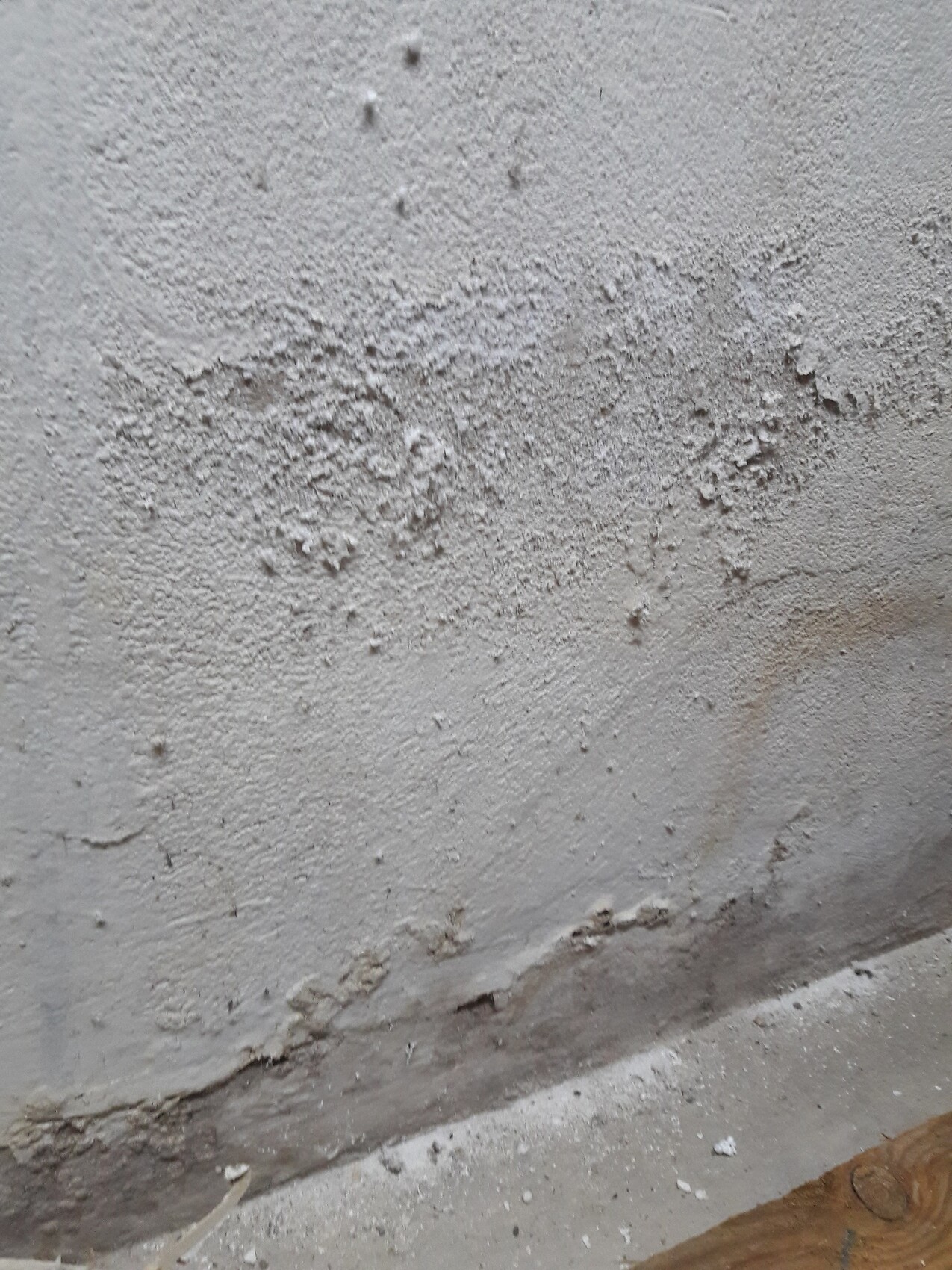 Graue, raue Putzwand mit Abplatzungen; Boden staubig mit Holzstück am Rand.
