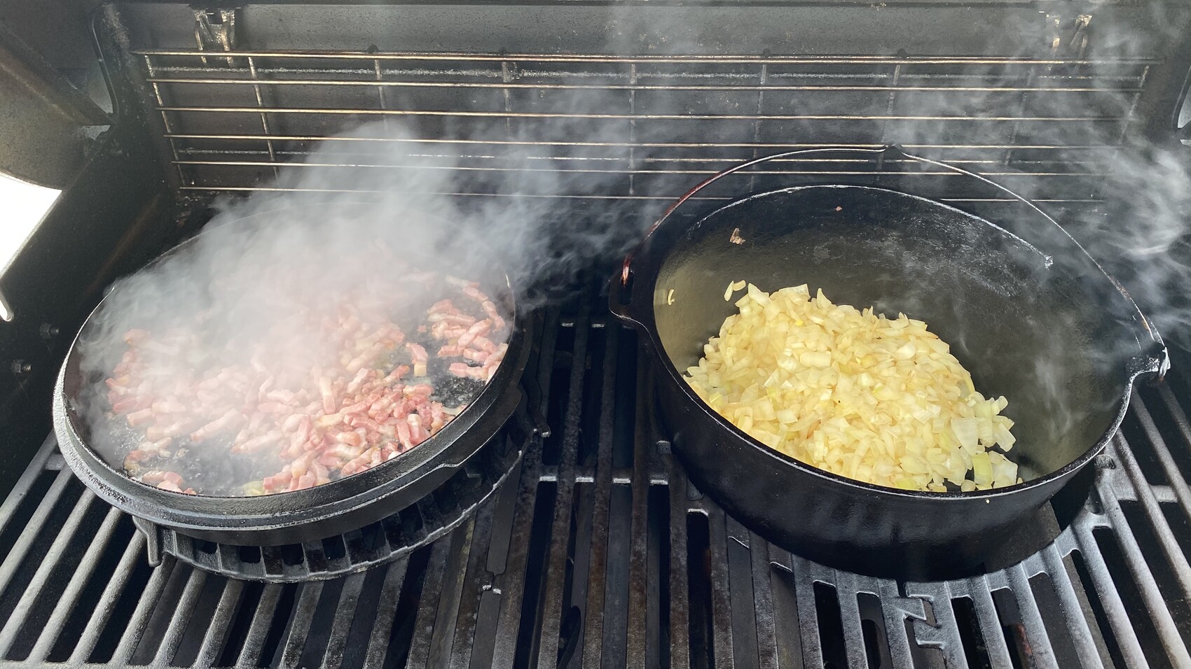 Zwei gusseiserne Pfannen auf dem Grill; links rohes gewürfeltes Fleisch, rechts Zwiebelwürfel, Rauch.