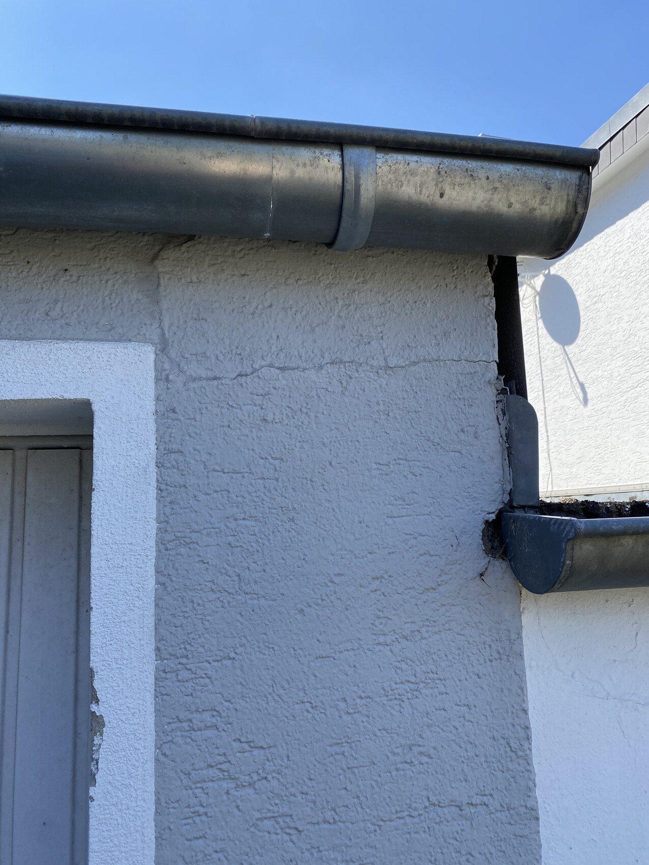 Dunkle Metall-Dachrinne an rauer Aussenwand, Ecke mit Riss; Satellitenschüssel-Schatten.
