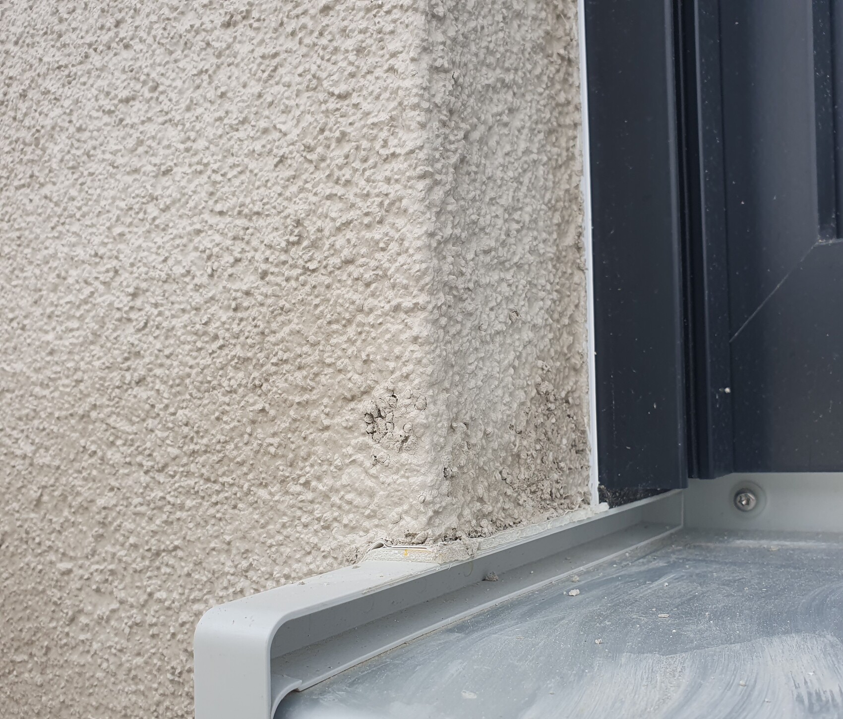 Außenwand mit grobem Putz, unten kleine Abplatzungen; daneben dunkler Fensterrahmen.