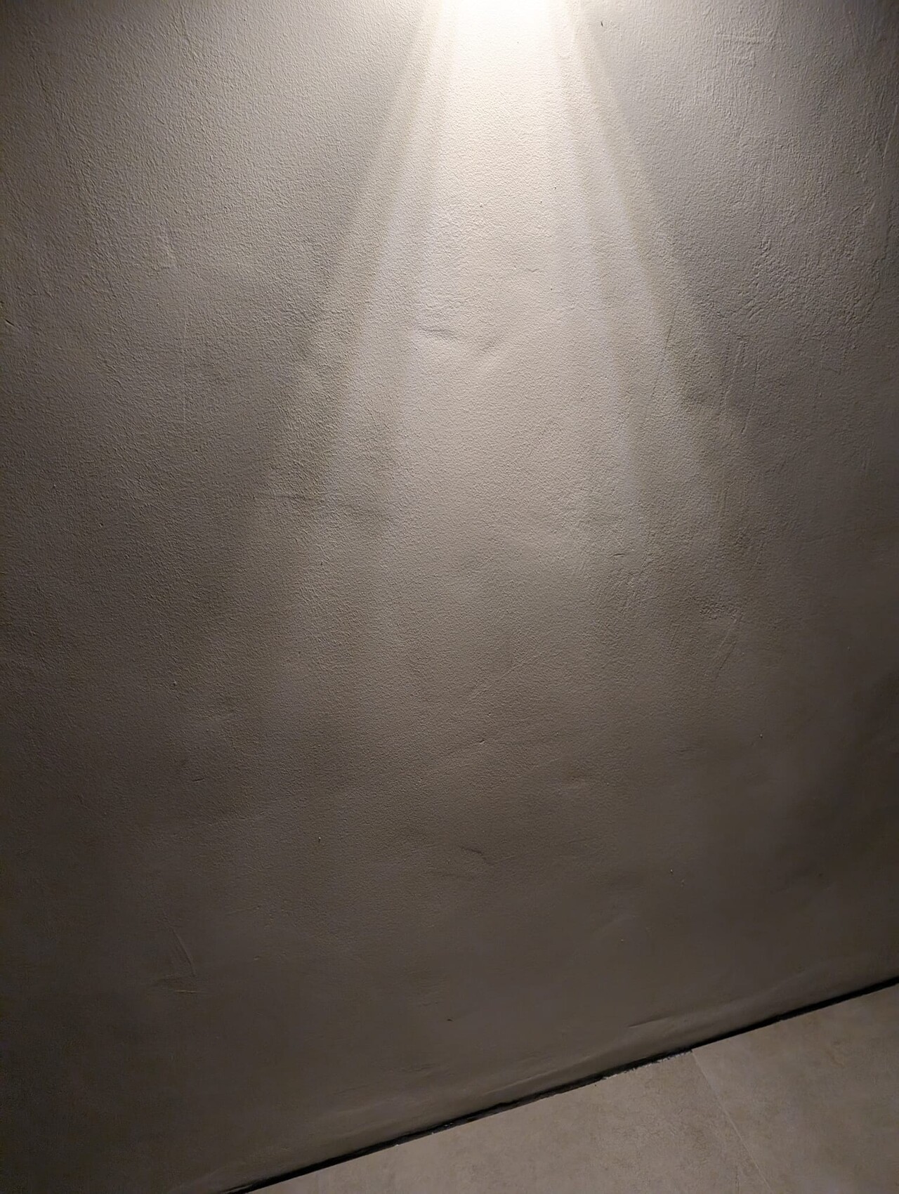 Grau verputzte Innenwand mit Lichtstrahl von oben, Bodenfliesen unten zu sehen.