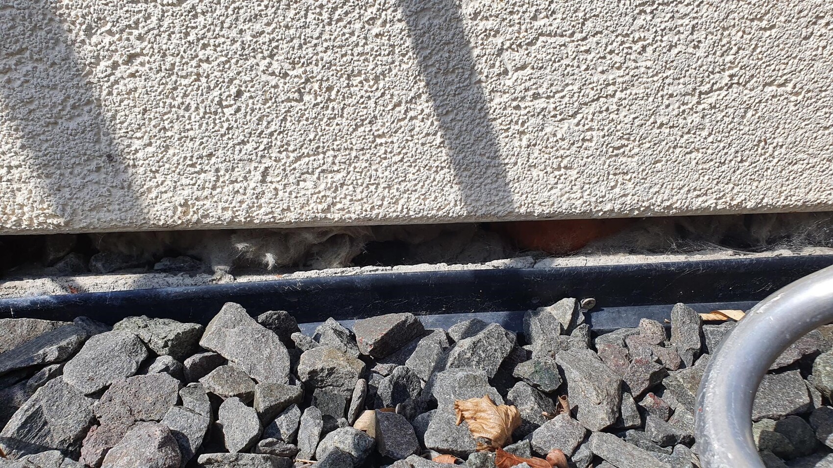 Raue beige Stuckwand, Schatten; Kieselsteine im Vordergrund, schwarze Rinne, Metallbogen rechts.