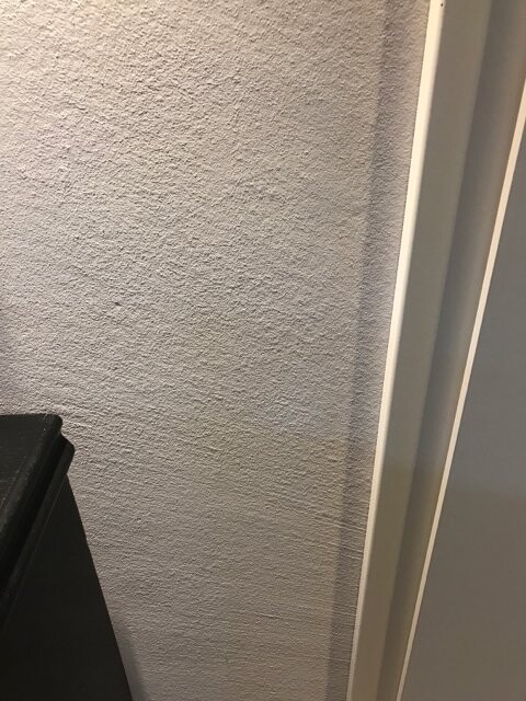 Beigefarbene, raue Wandfläche mit Struktur; rechts Türrahmen, links schwarzes Möbelteil.