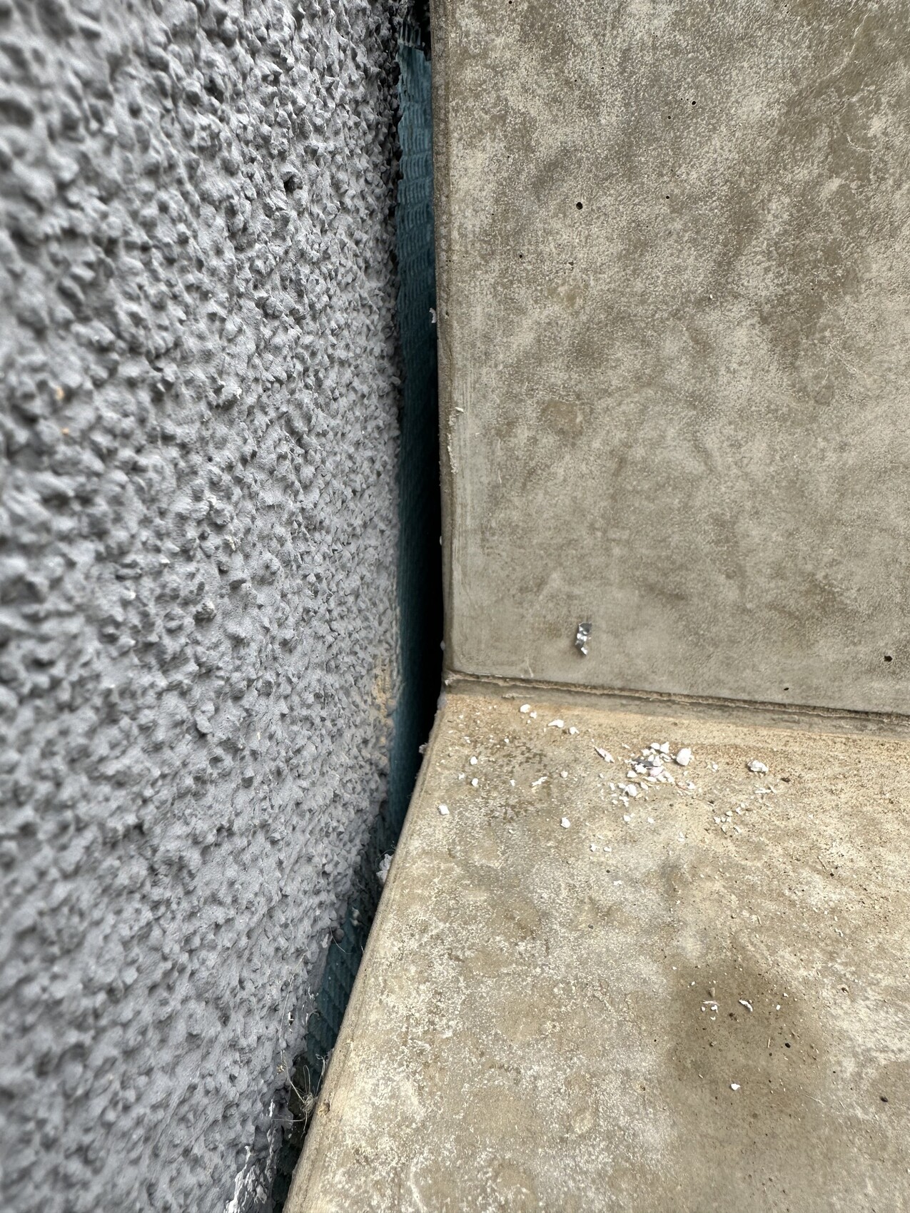 Ecke zwischen rauem Putz links und glattem Beton rechts; staubiger Boden.