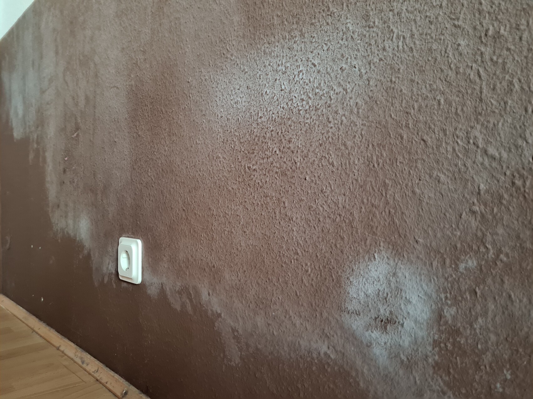 Rauhe braune Wand mit Struktur; weiße Steckdose links unten, Holzbodenleiste sichtbar.