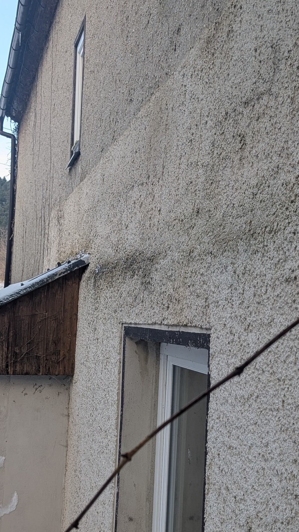 Außenwand mit Rauhputz, Fenster, Holzkonstruktion und Drahtzaun im Vordergrund.