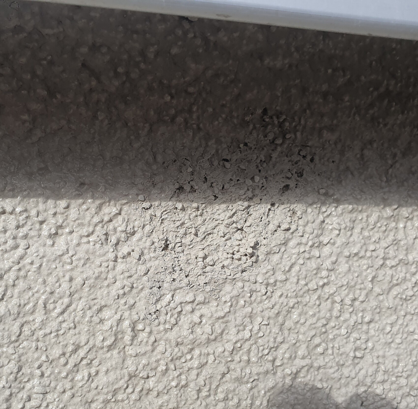 Beigefarbene Putzwand mit grober Struktur und dunkler Schattenlinie unter einer Metallkante.