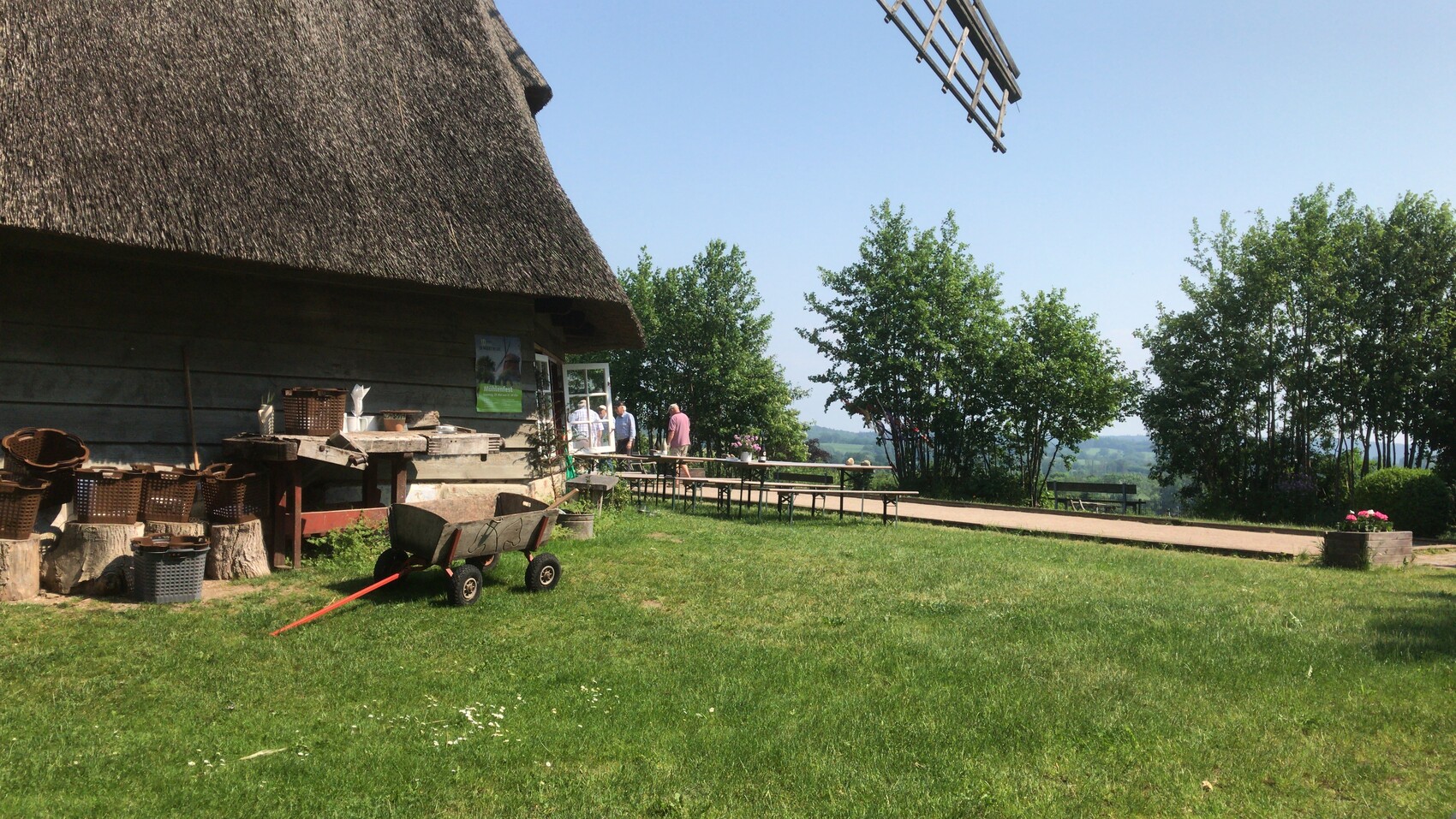 Reetdachhaus mit Vorderseite aus Holz; Schubkarre, Körbe, Tische davor, grüne Landschaft
