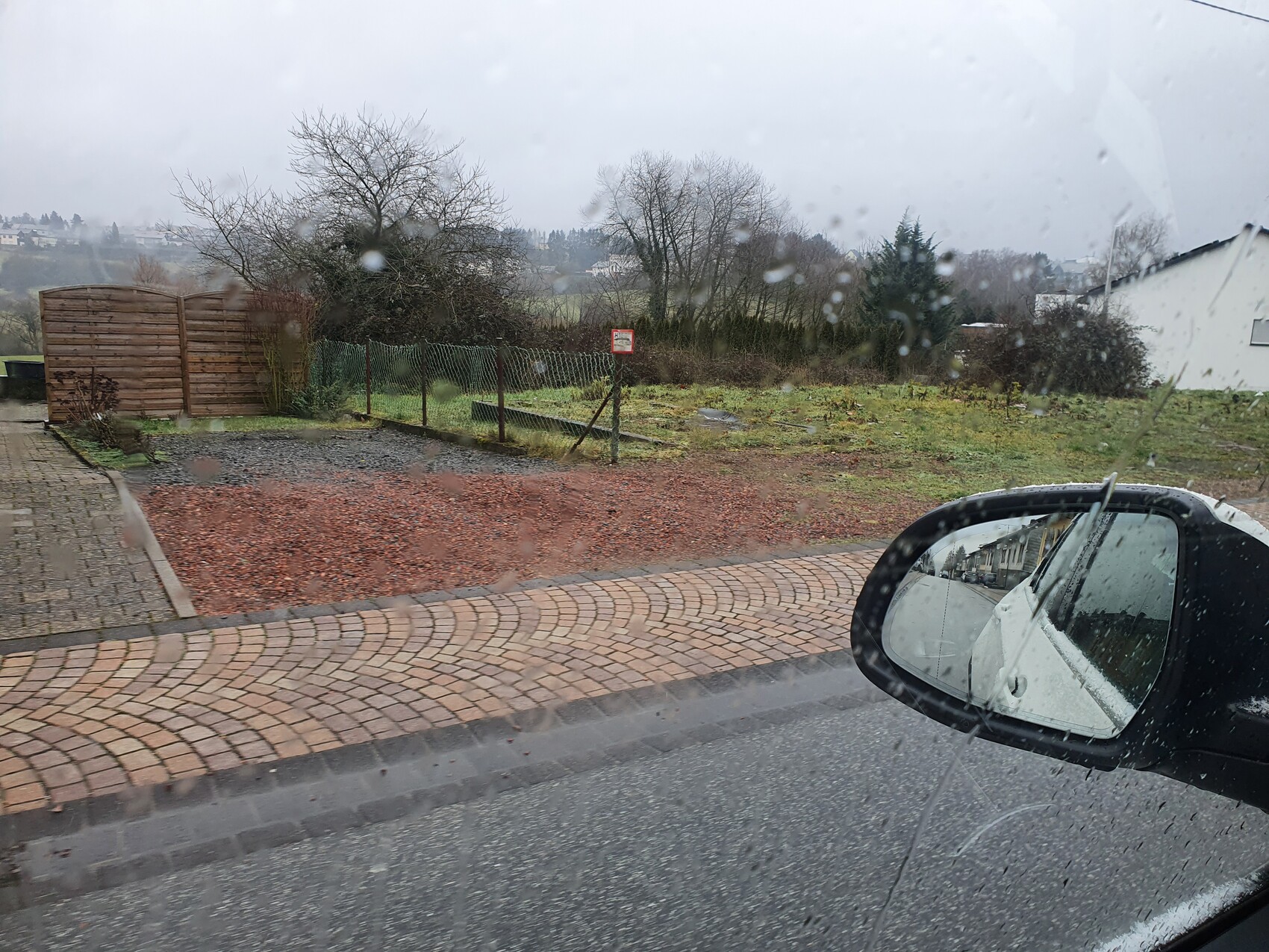 Blick aus Auto bei Regen: nasse Scheibe, Kopfsteinpflaster, Gartenzaun, Spiegel zeigt Straßenzug.