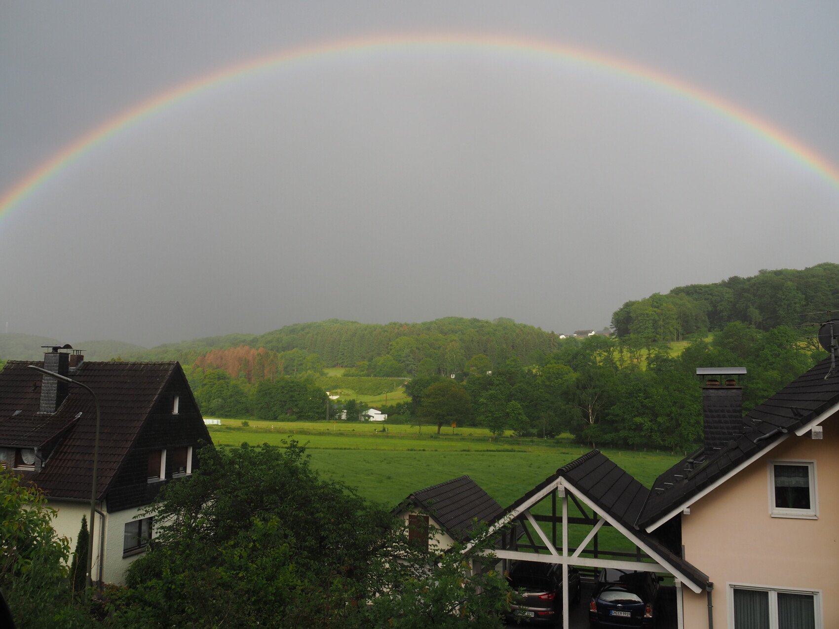 Regenbogen spannt sich über grüne Hügellandschaft mit Häusern im Vordergrund.