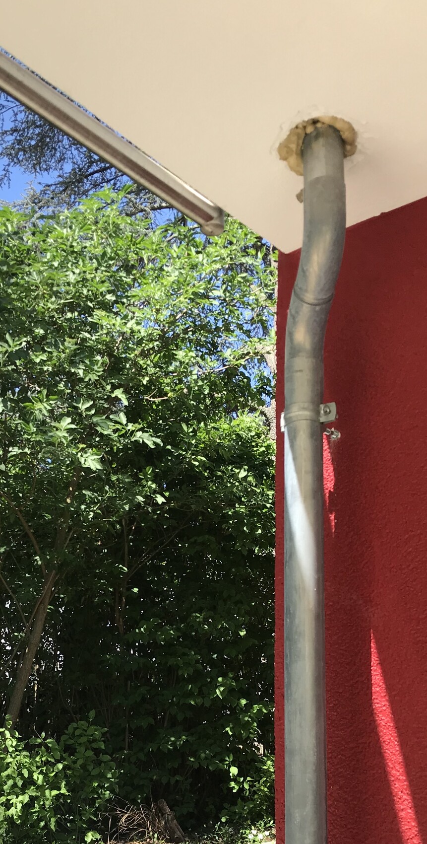 Außenbereich: senkrechtes Metall-Fallrohr an roter Wand, unter weißer Decke, grüne Hecke und blauer Himmel.