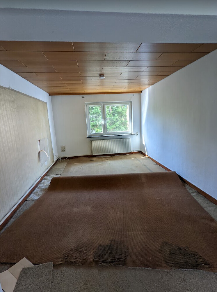 Schmaler Dachbodenraum mit Holzdecke, weißen Wänden und braunem Teppich; Fenster mit Heizkörper.