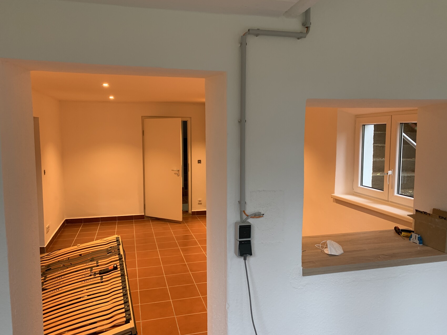 Wohnraum mit braunen Fliesen, Tür offen, Bettgestell vorn, Küchenfenster rechts.
