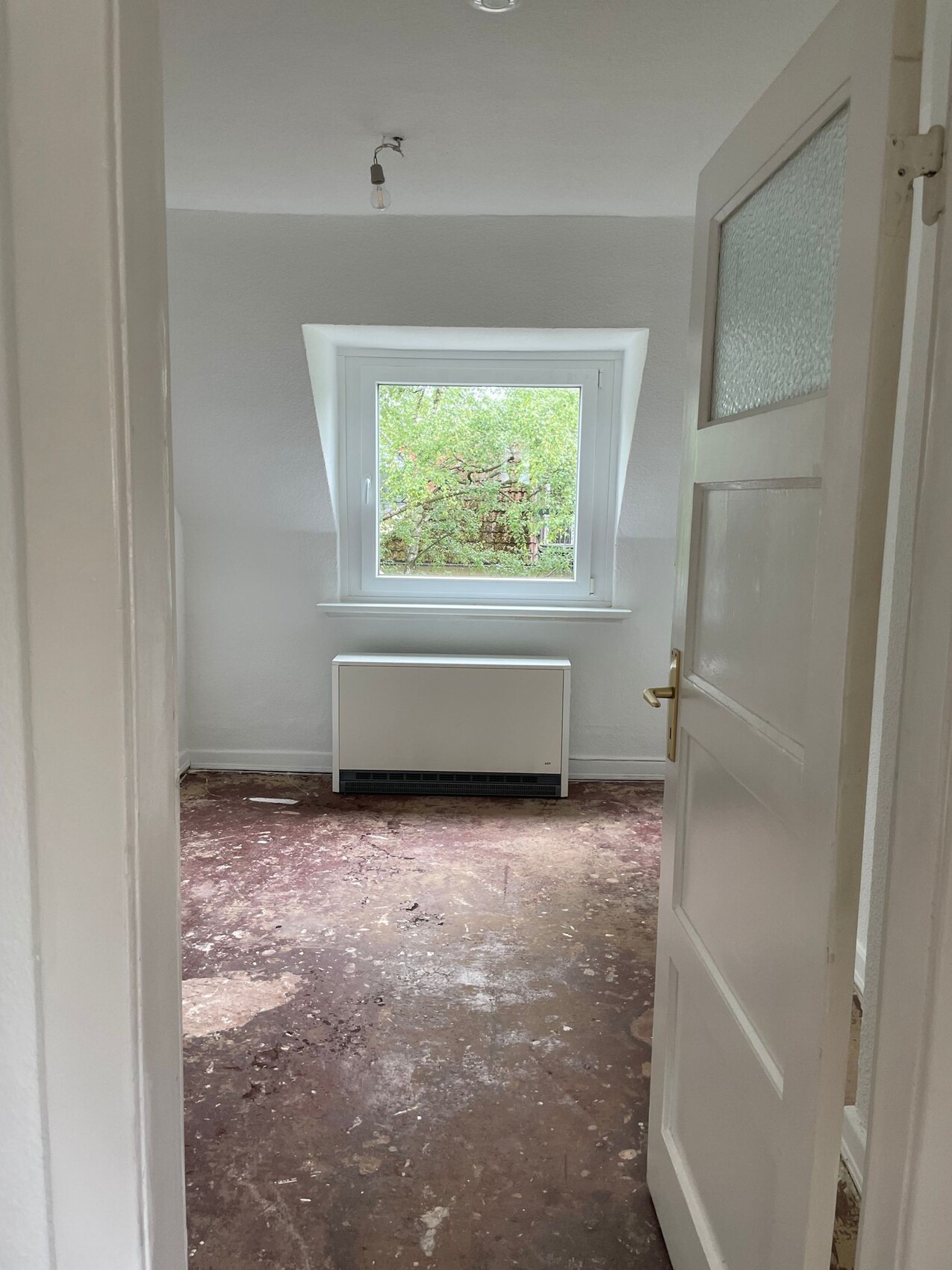 Kleines, helles Dachgeschoss-Zimmer mit Fenster; darunter Heizgerät; offene Tür; Boden stark abgenutzt.