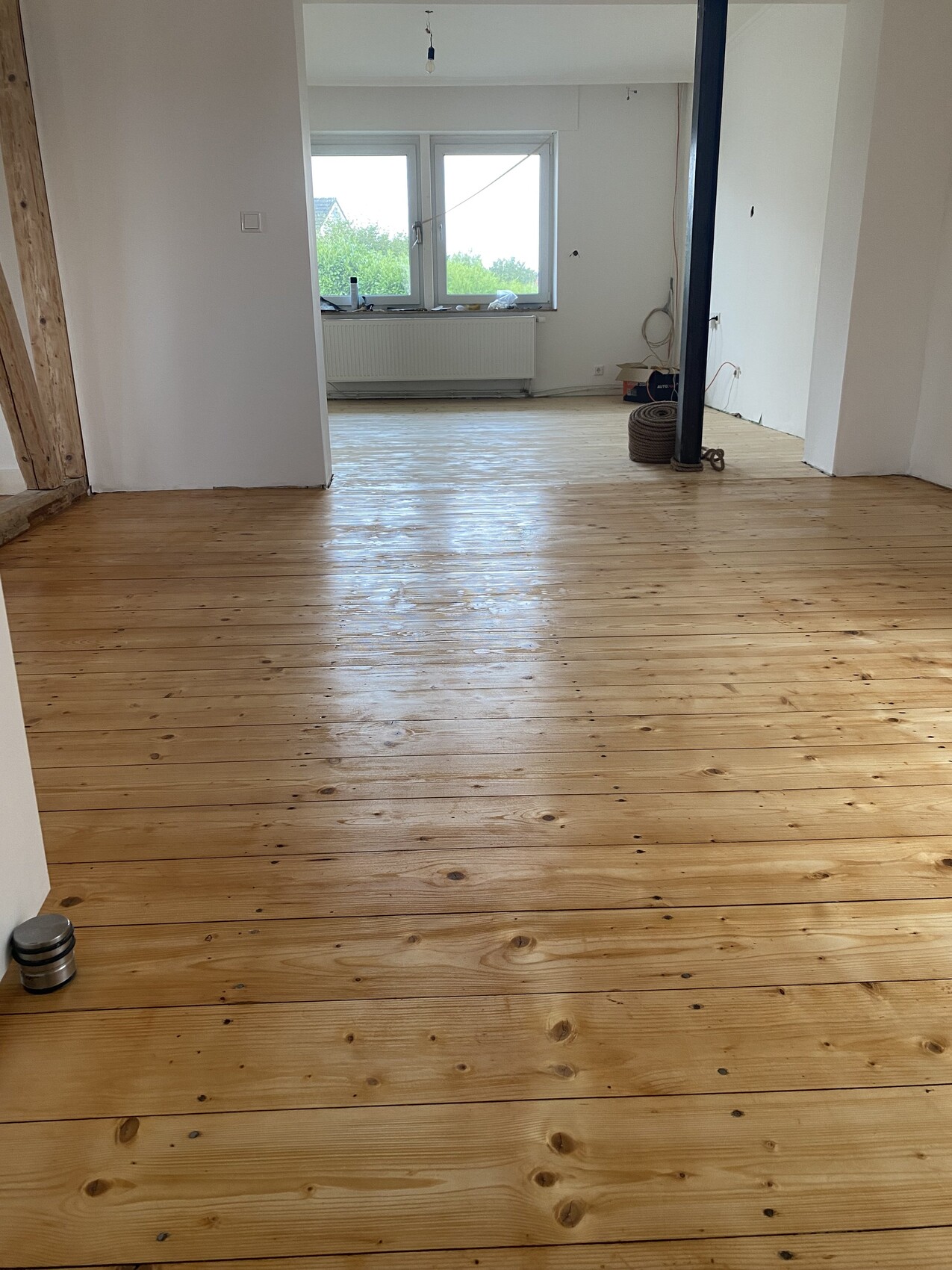 Leeres Zimmer in Renovierungszustand mit hellem Holzboden, Fensterfront, Heizkörper und Metallstütze.