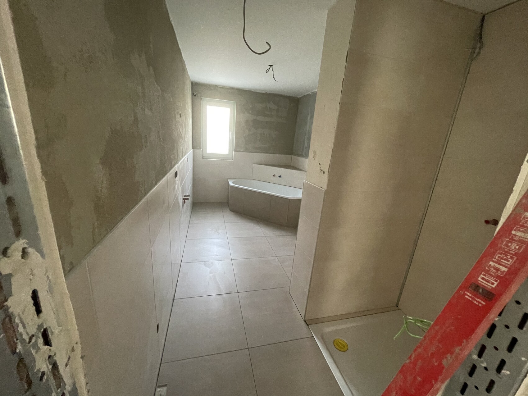 Unfertiges Badezimmer unter Renovierung: Badewanne, weiße Fliesen, Fenster, lose Kabel.