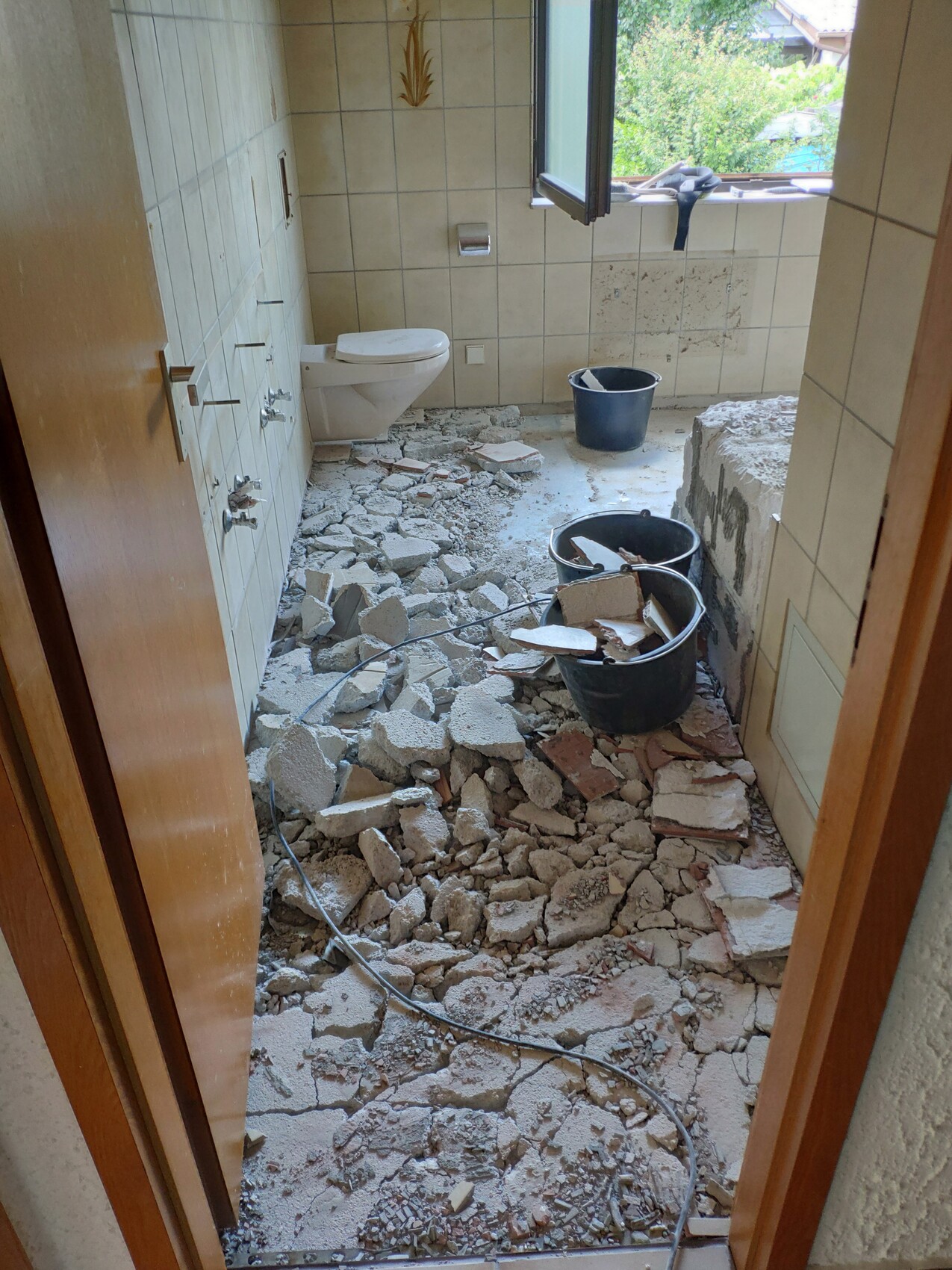 Badezimmer im Umbau: Boden voller Steine und Staub, Toilette, Eimer und offenes Fenster.