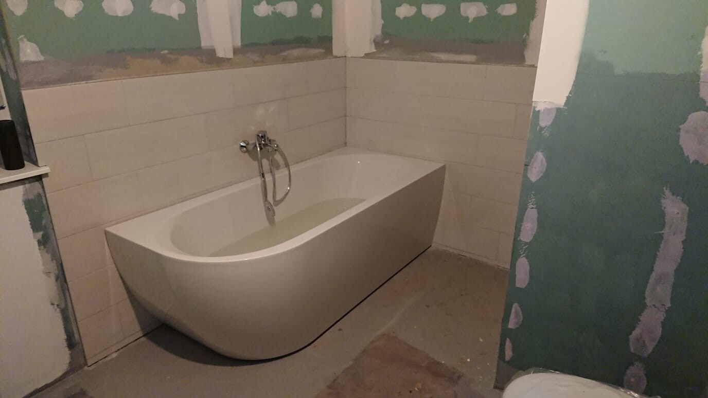Freistehende weiße Badewanne vor unvollendeten Fliesen und grünen Wandflächen im Bad.