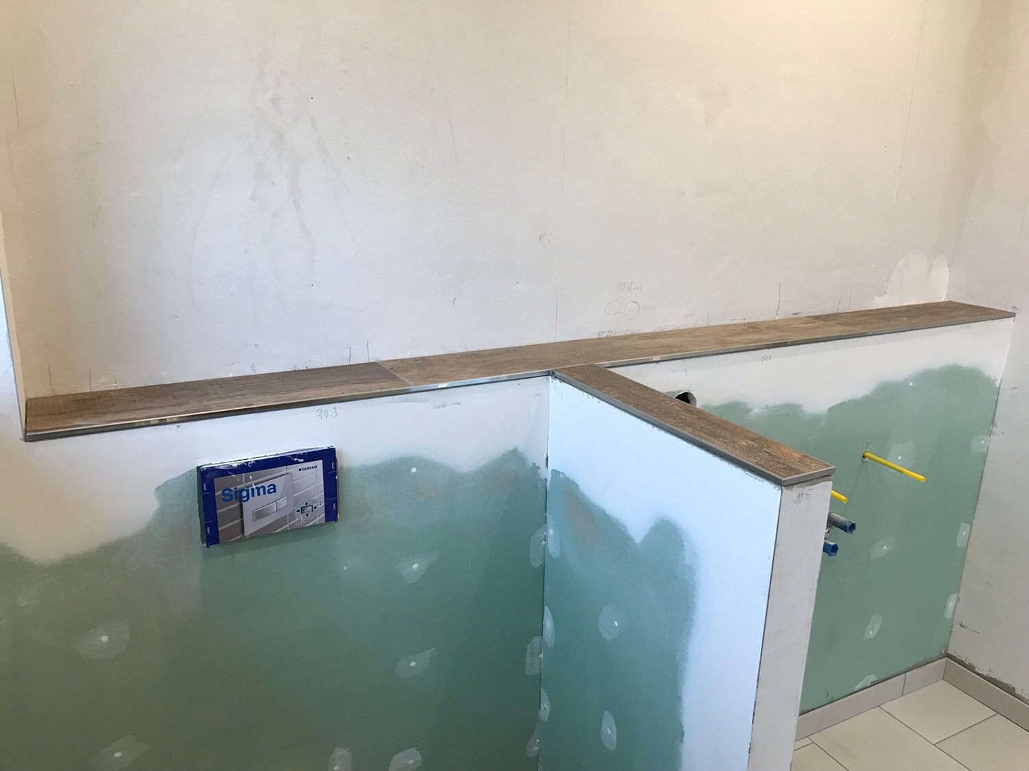 Baustelle: grüne Trockenbauwand, Holzablage, Rohrleitungen rechts, Sigma-Verpackung an Wand.