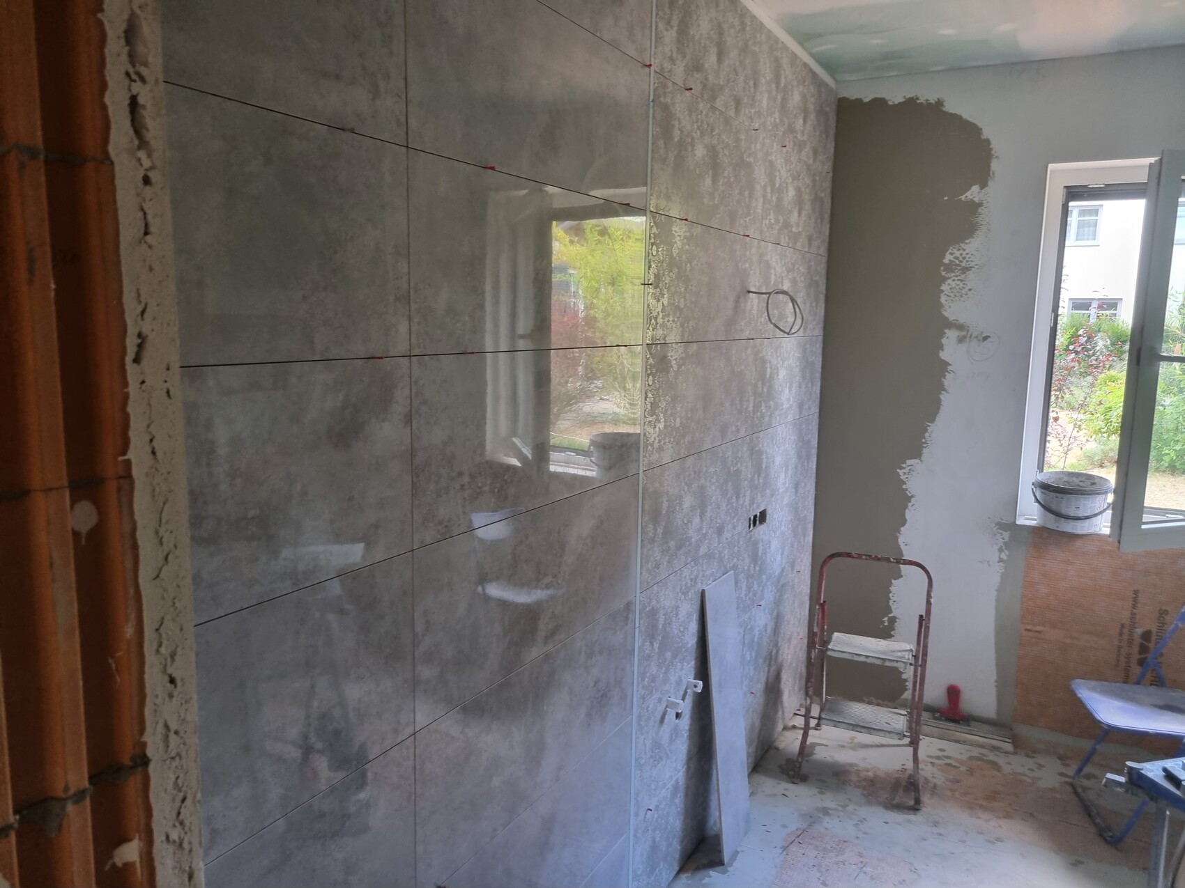 Baustelle: neue graue Fliesenwand wird verlegt; Leiter, Eimer, offenes Fenster.