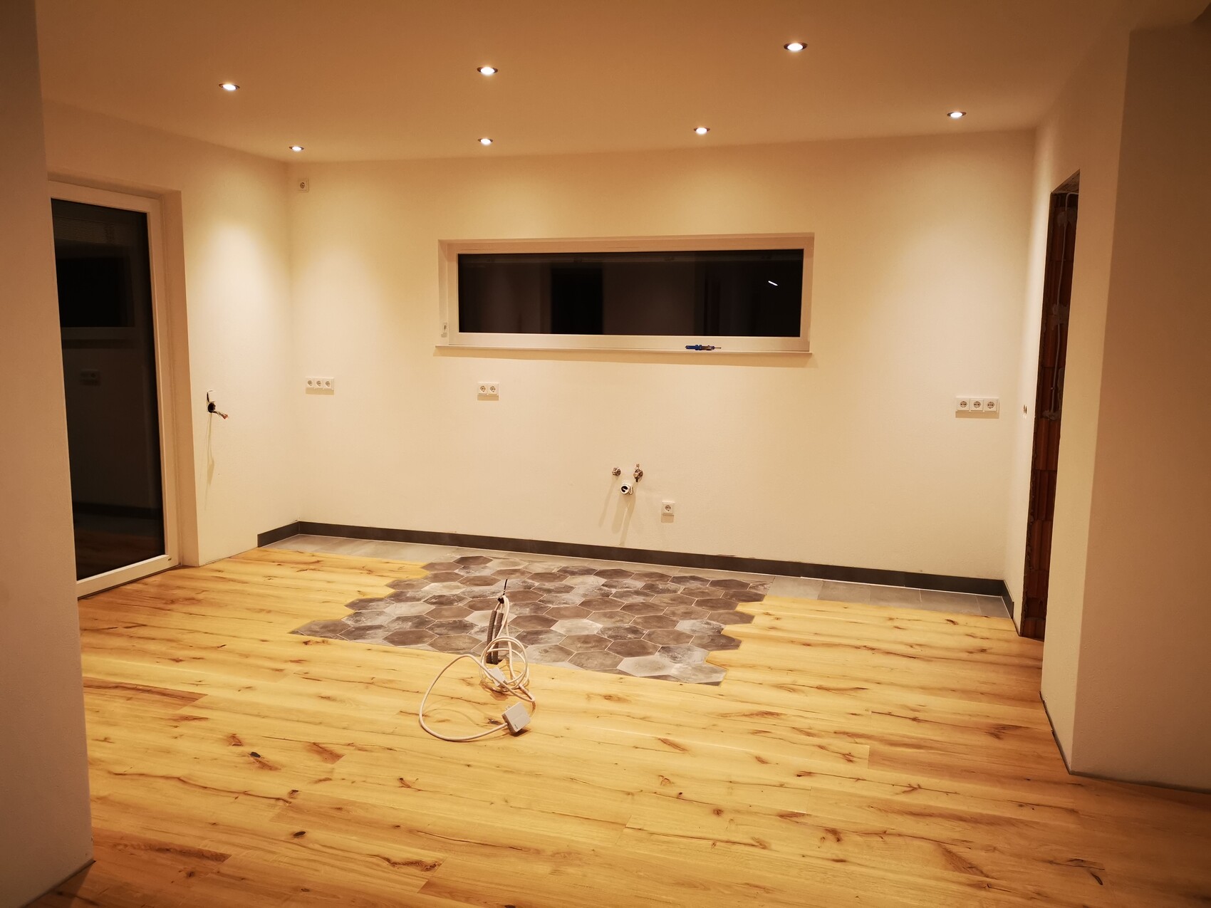 Heller Renovierungsraum mit Holzboden, zentrale graue Fliesenfläche und lose Kabel.