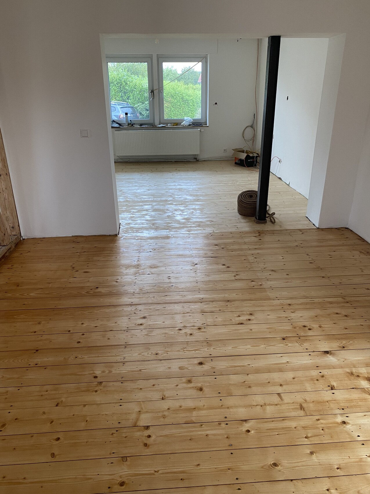 Heller Renovierungsraum mit Holzfußboden, offenem Durchgang und Fenster mit Heizkörper darunter.