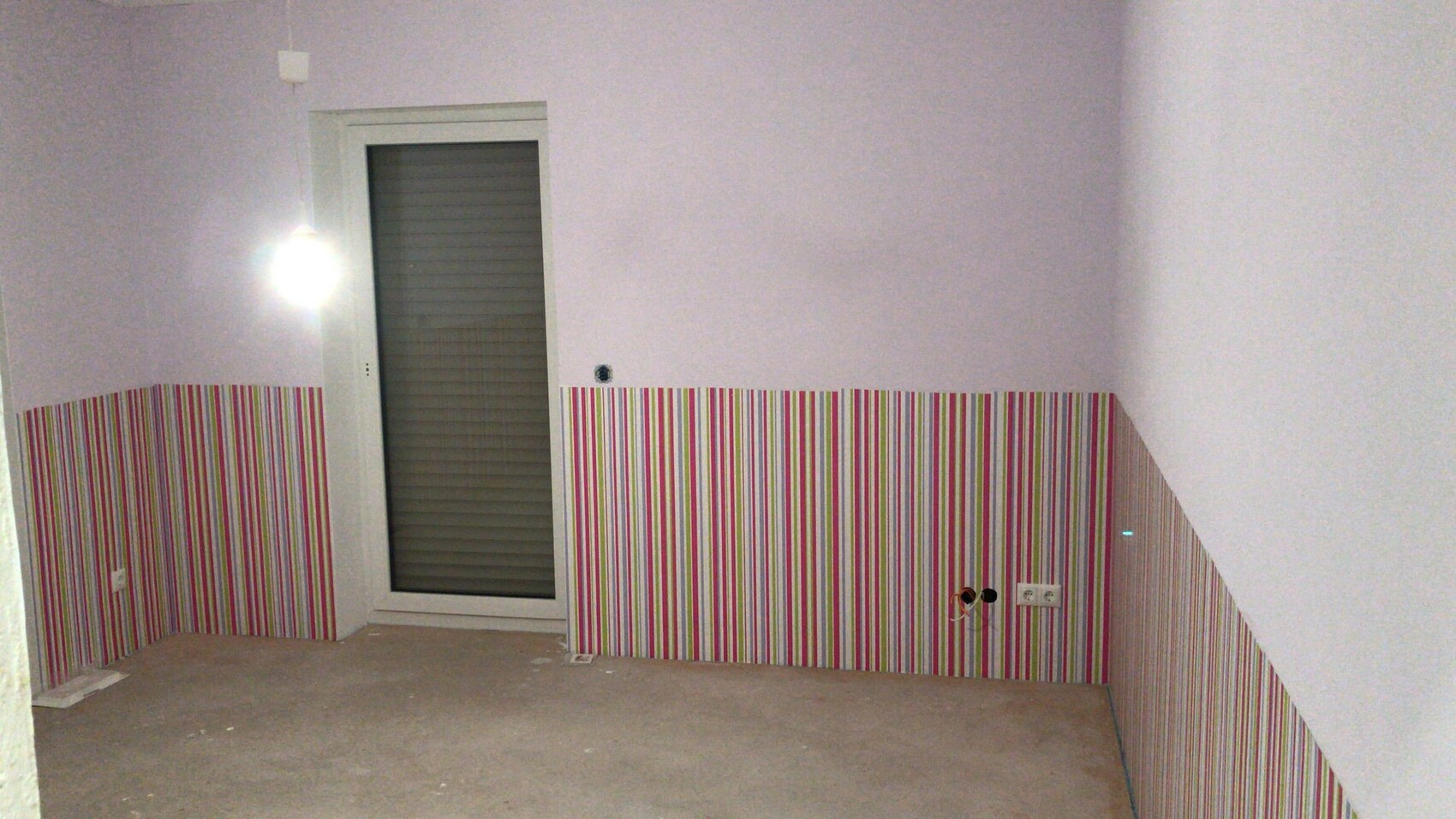 Kleines Zimmer mit pastellvioletter Oberwand, unten bunte Streifenverkleidung, Tür mit Rollladen.