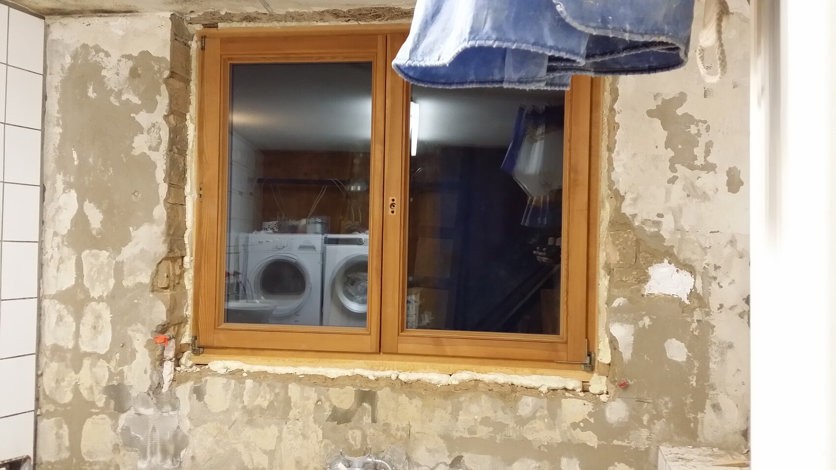 Holzfenster in renovierter, rauer Wand; hinter Glas stehen zwei Frontlader-Waschmaschinen.
