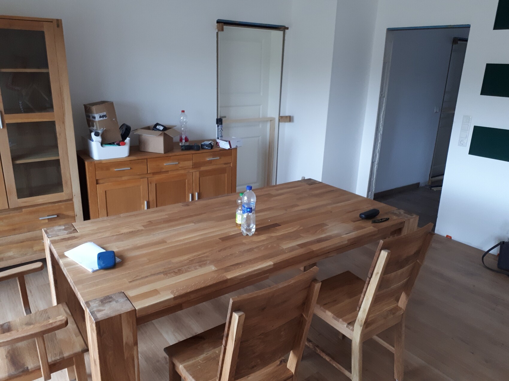 Großer Holztisch mit Stühlen in unaufgeräumtem Esszimmer; Kartons, Flasche auf der Anrichte.