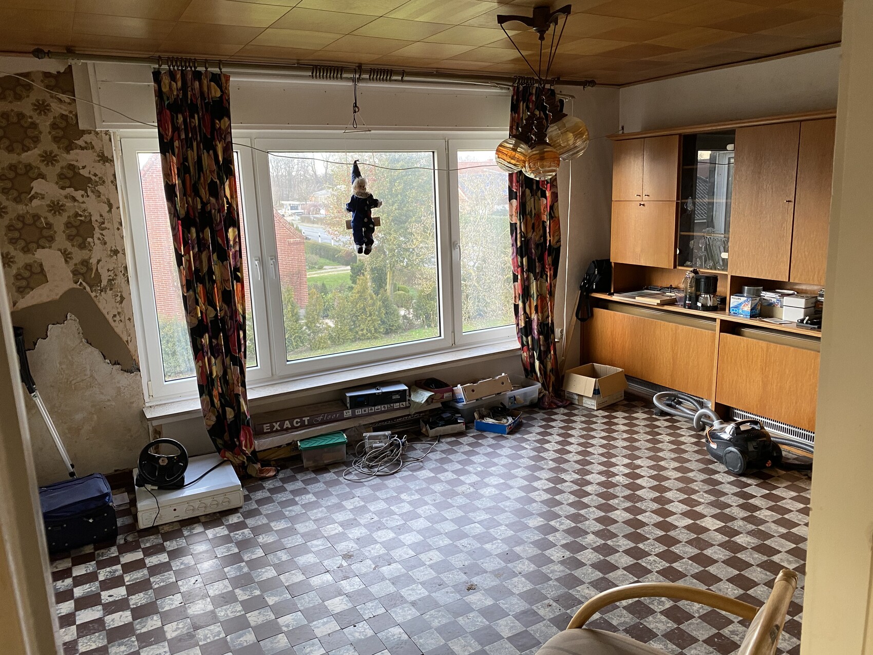 Wohnzimmer mit großen Fenstern, bunten Vorhängen, einem hängenden Plüschtier und Holzschränken.