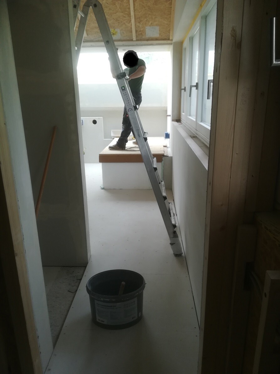 Mann steht auf einer Leiter in einem Bauinnenraum und arbeitet an der Decke; Eimer steht am Boden