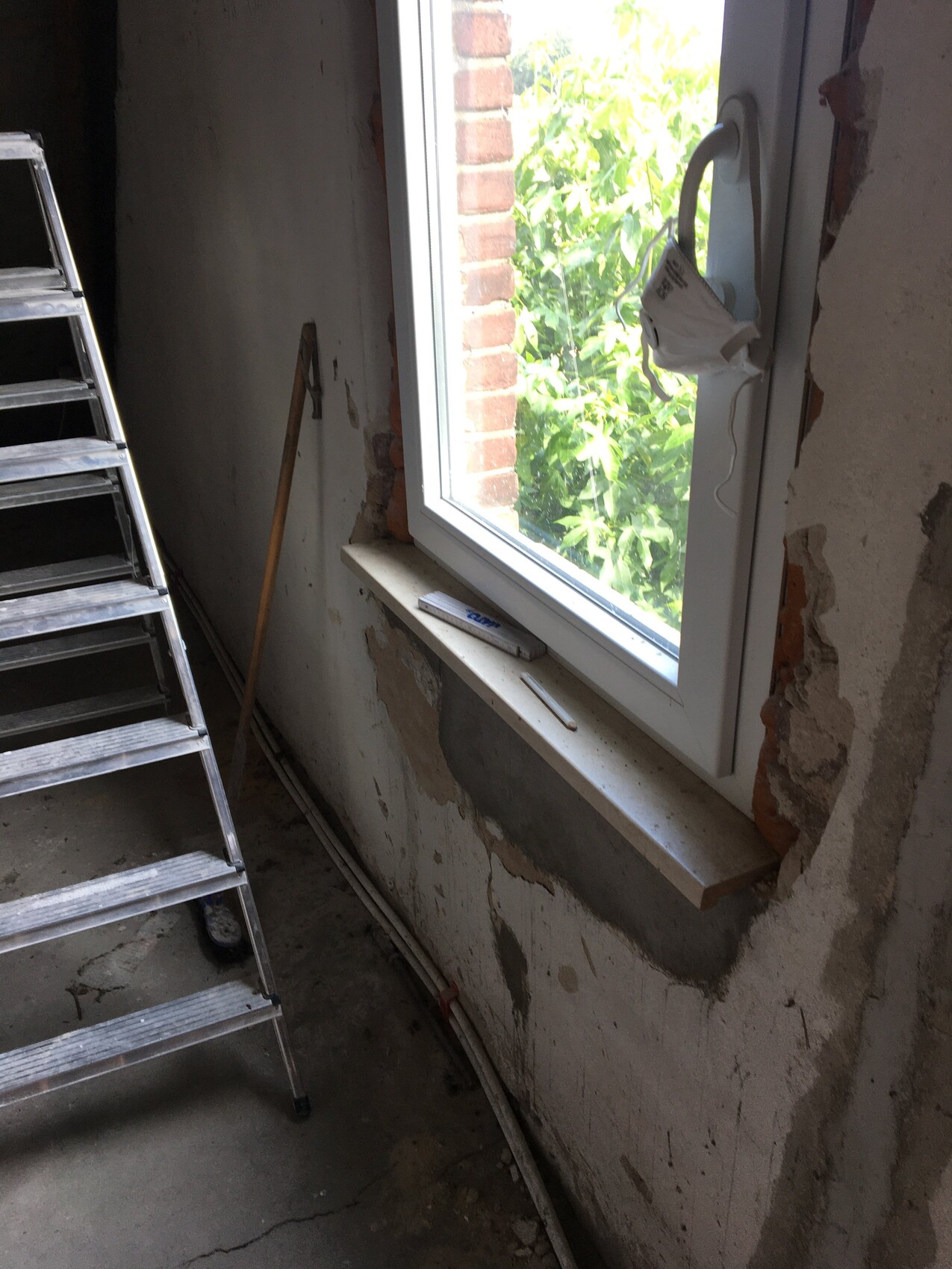 Unfertiger Innenraum einer Baustelle: Fensterrahmen, Leiter links, Putzreste, Maske am Fenstergriff
