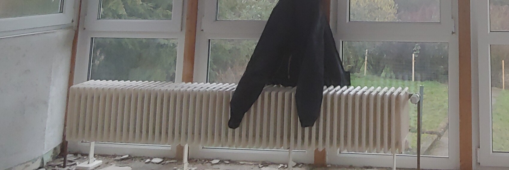 Schwarze Jacke haengt ueber dem Heizkoerper vor grossen Fenstern in einem unaufgeraeumten Raum.