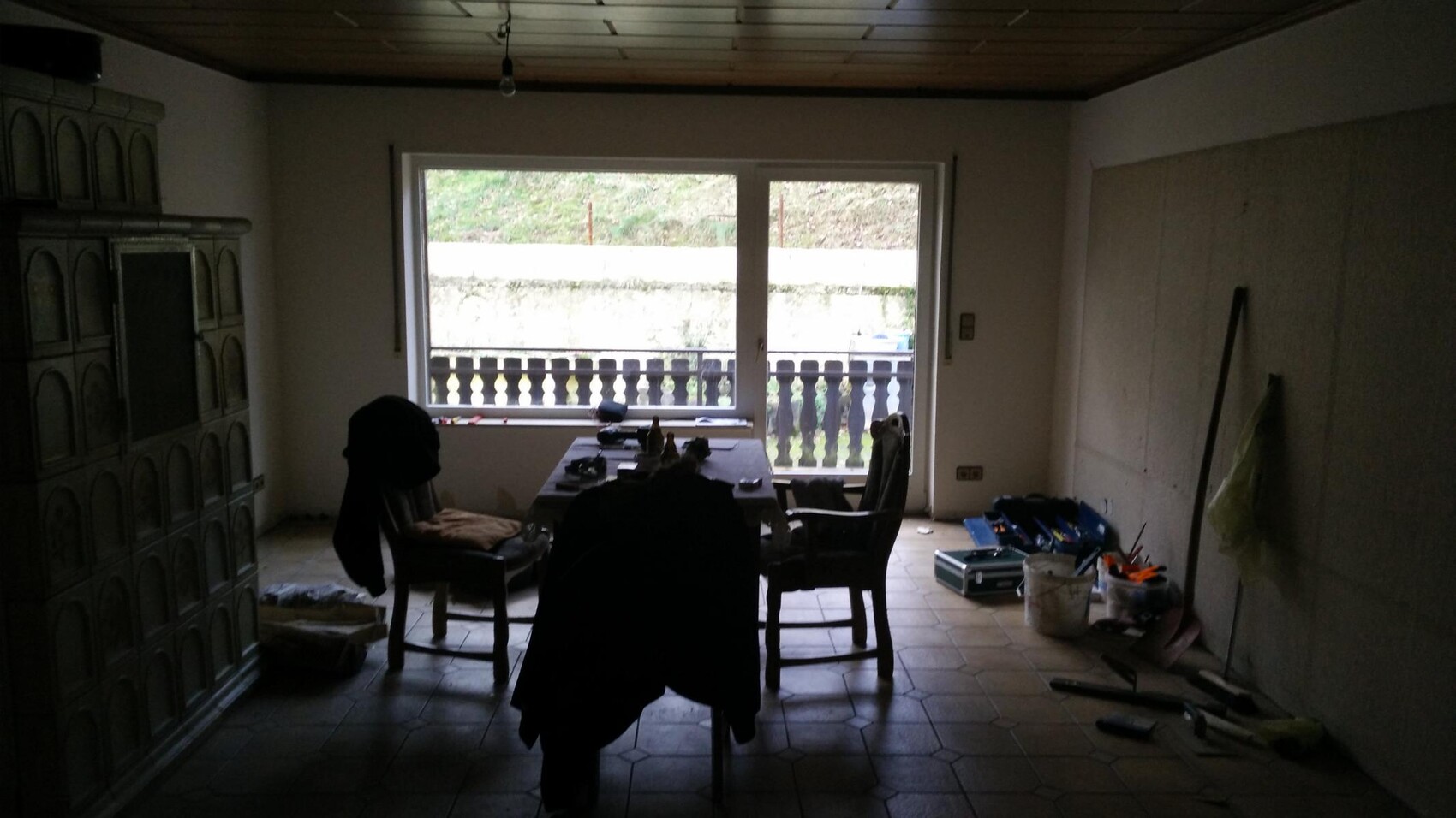 Dunkler Kellerraum mit Esstisch, Stühlen und Bauwerkzeugen; Balkonblick durch große Fenster