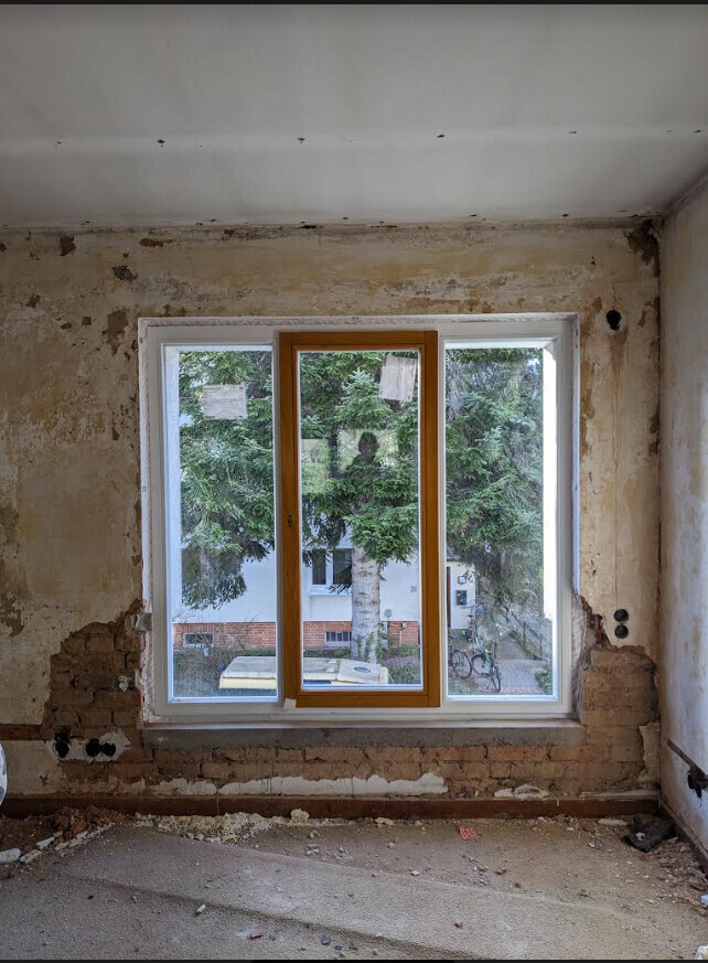 Rohbauzimmer mit großem Fensterrahmen aus Holz; bröckelnde Wand, Blick nach außen auf Bäume.