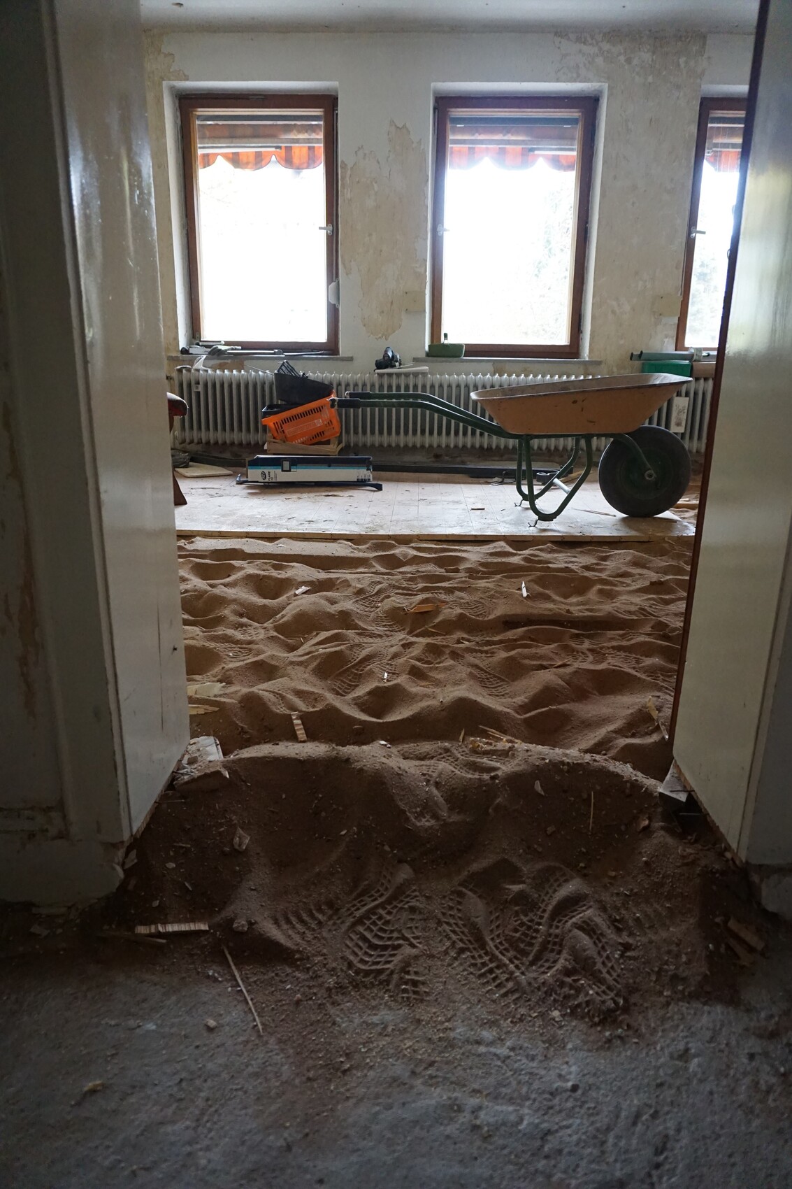 Raum im Renovierungszustand mit Sandboden, Schubkarre, Baumaterial und drei Fenster.