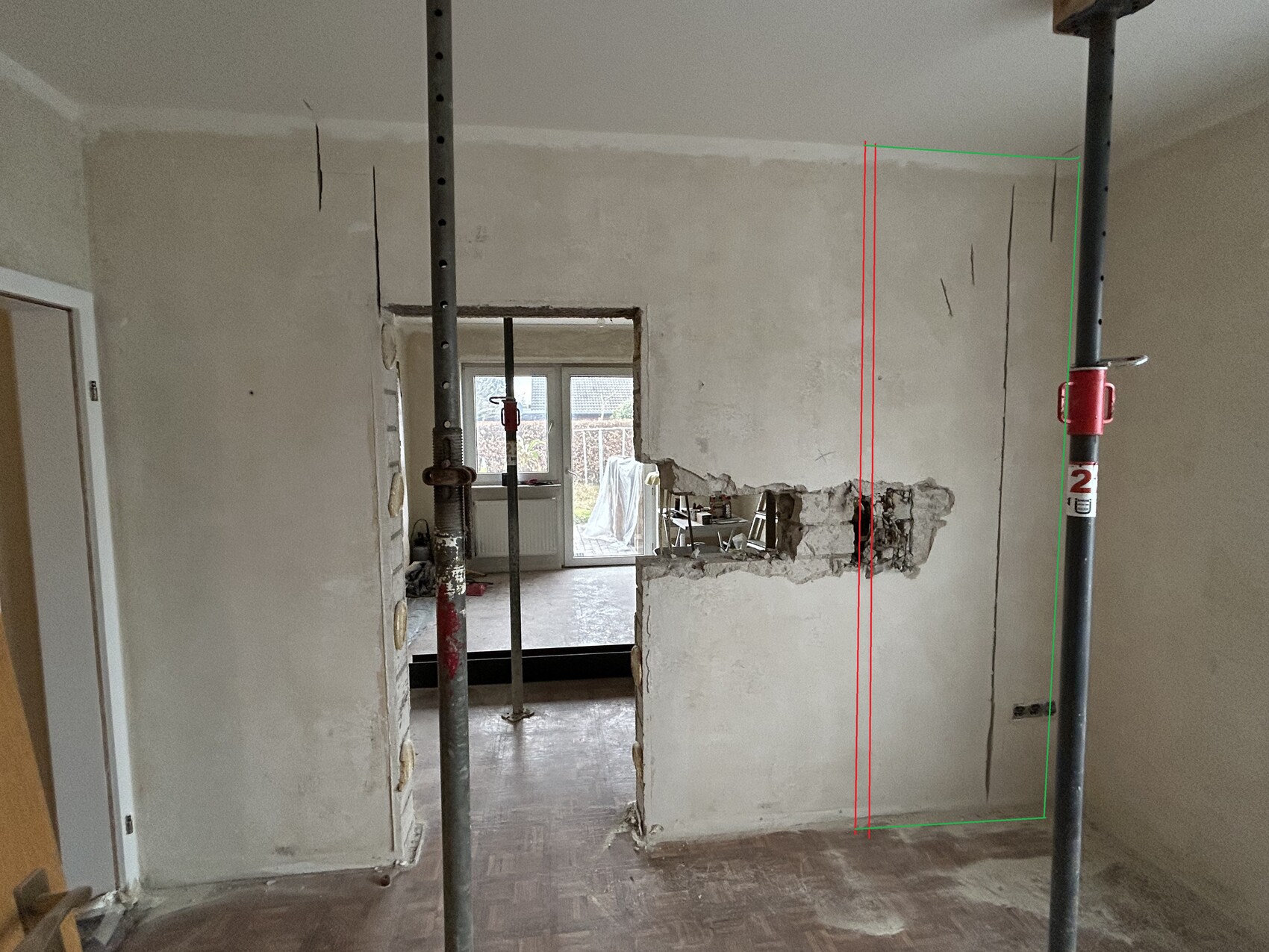 Baustellenraum: Stahlstützen, beschädigte Wand mit Loch, Blick in Nebenzimmer.