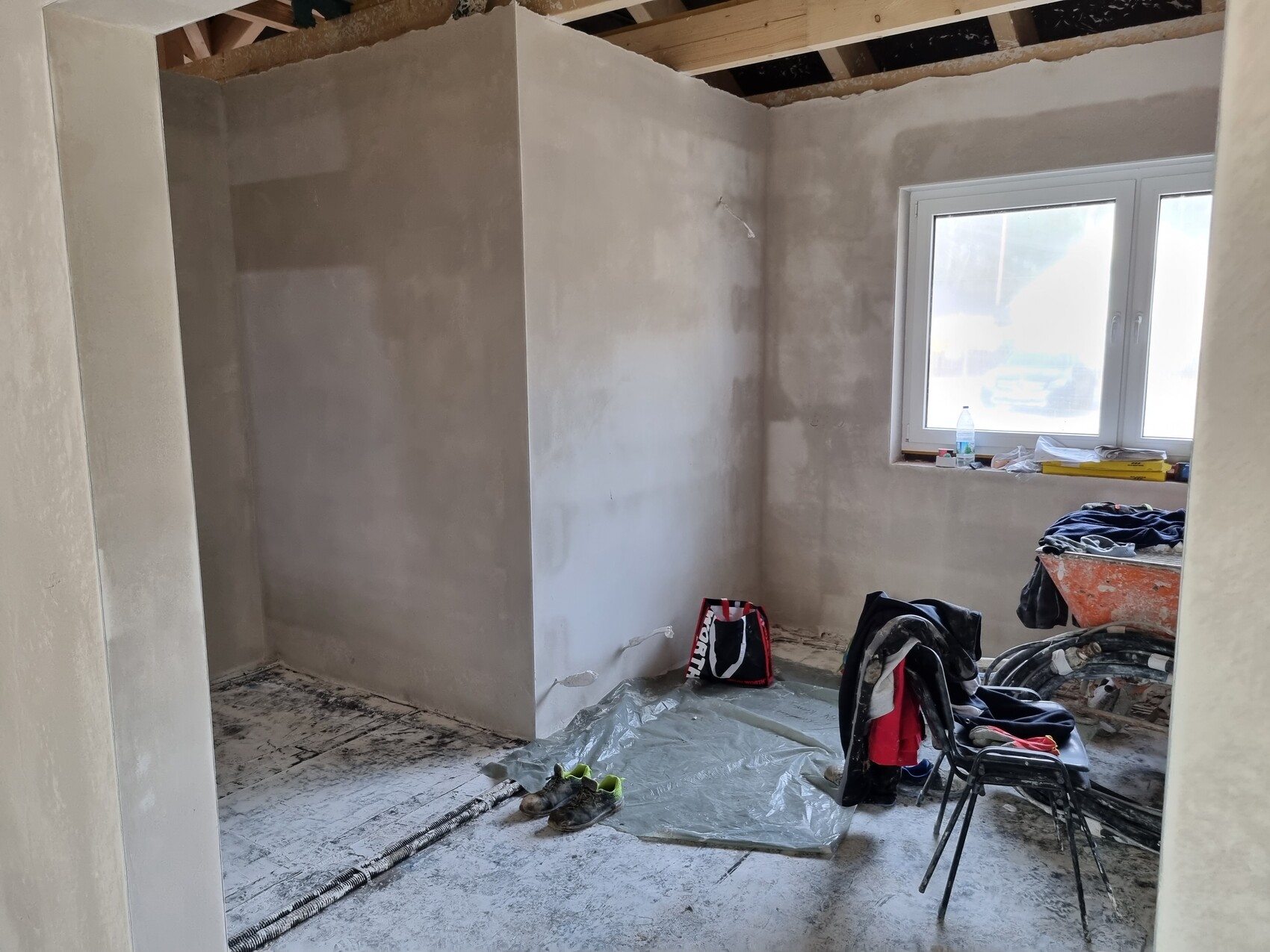Renovierungsraum mit unverputzten Wänden, Fenster, Abdeckfolie, Werkzeug und Kleidung.