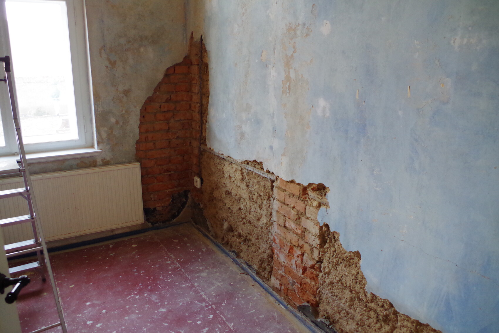 Renovierungsraum mit freiliegender Backsteinwand, blauem Putz, Leiter und Fenster.