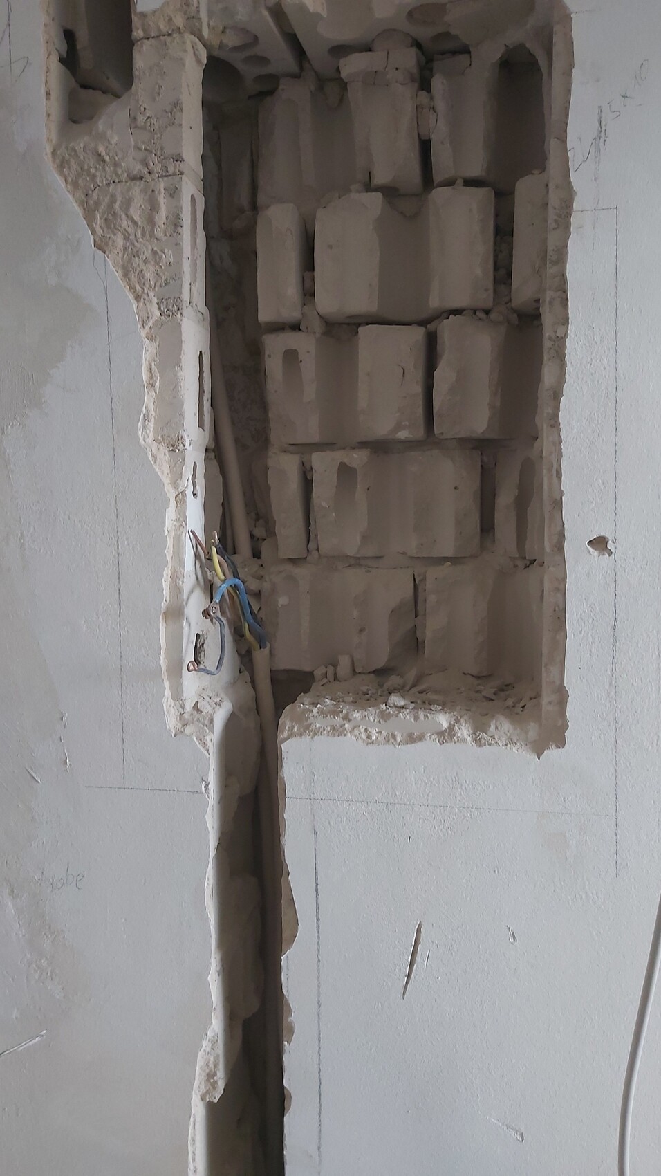 Durchbrochene Wand offenbart freiliegende Ziegelblöcke im Inneren; lose Kabel hängen seitlich.