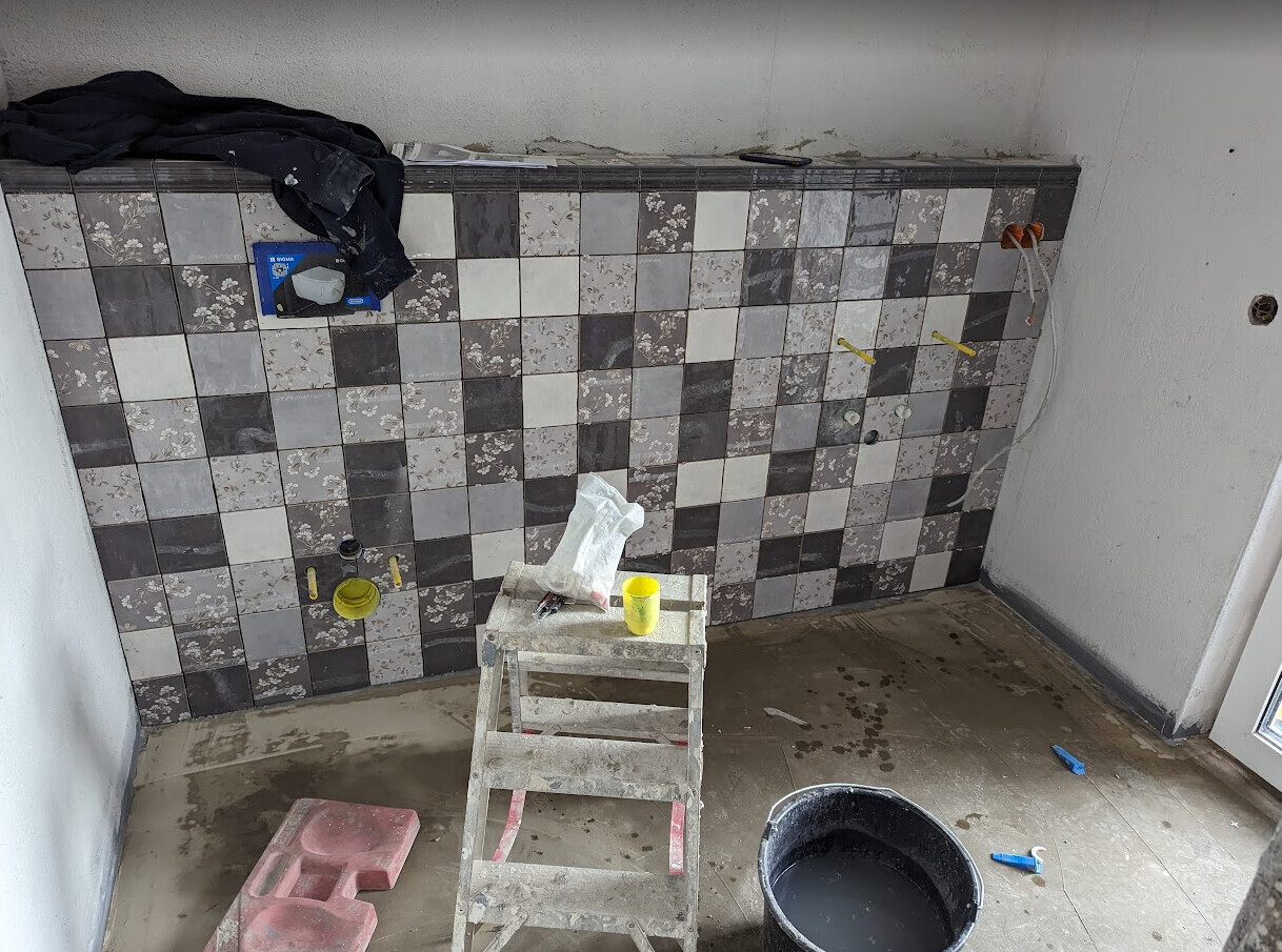 Renovierungsraum mit gemusterter Fliesenwand, Leiter, Eimer und Werkzeugen auf nassem Boden.