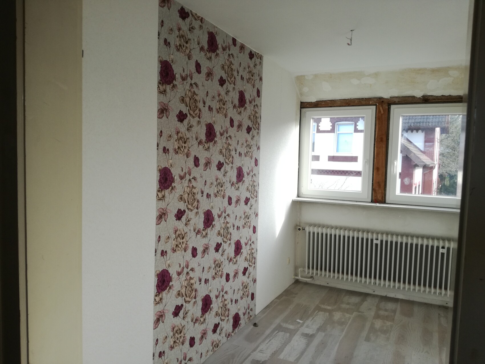 Kleines Zimmer mit floraler Wandtapete links, Fensterfront mit Heizkörper darunter.