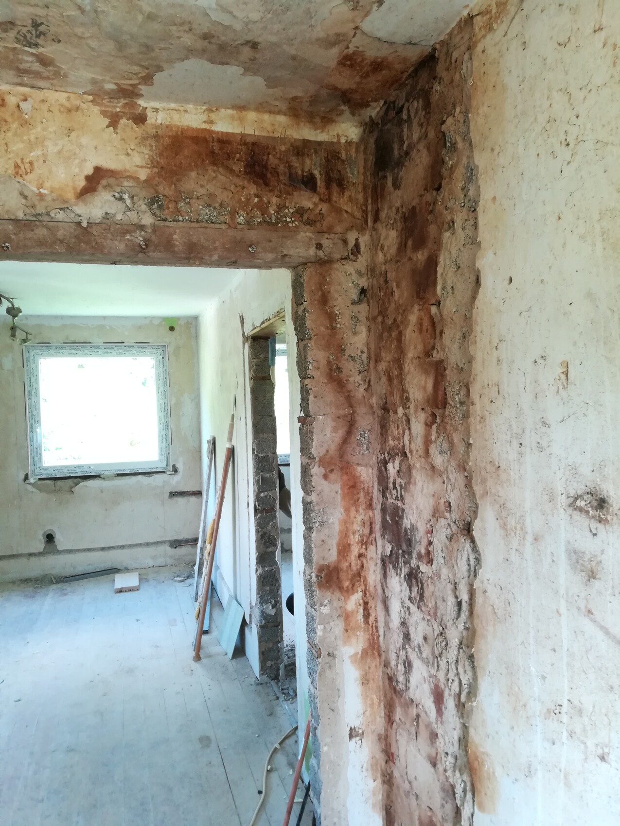 Renovierungsraum mit freigelegter Backsteinwand, Holzträgern, schäbigem Putz und Fensterlicht.