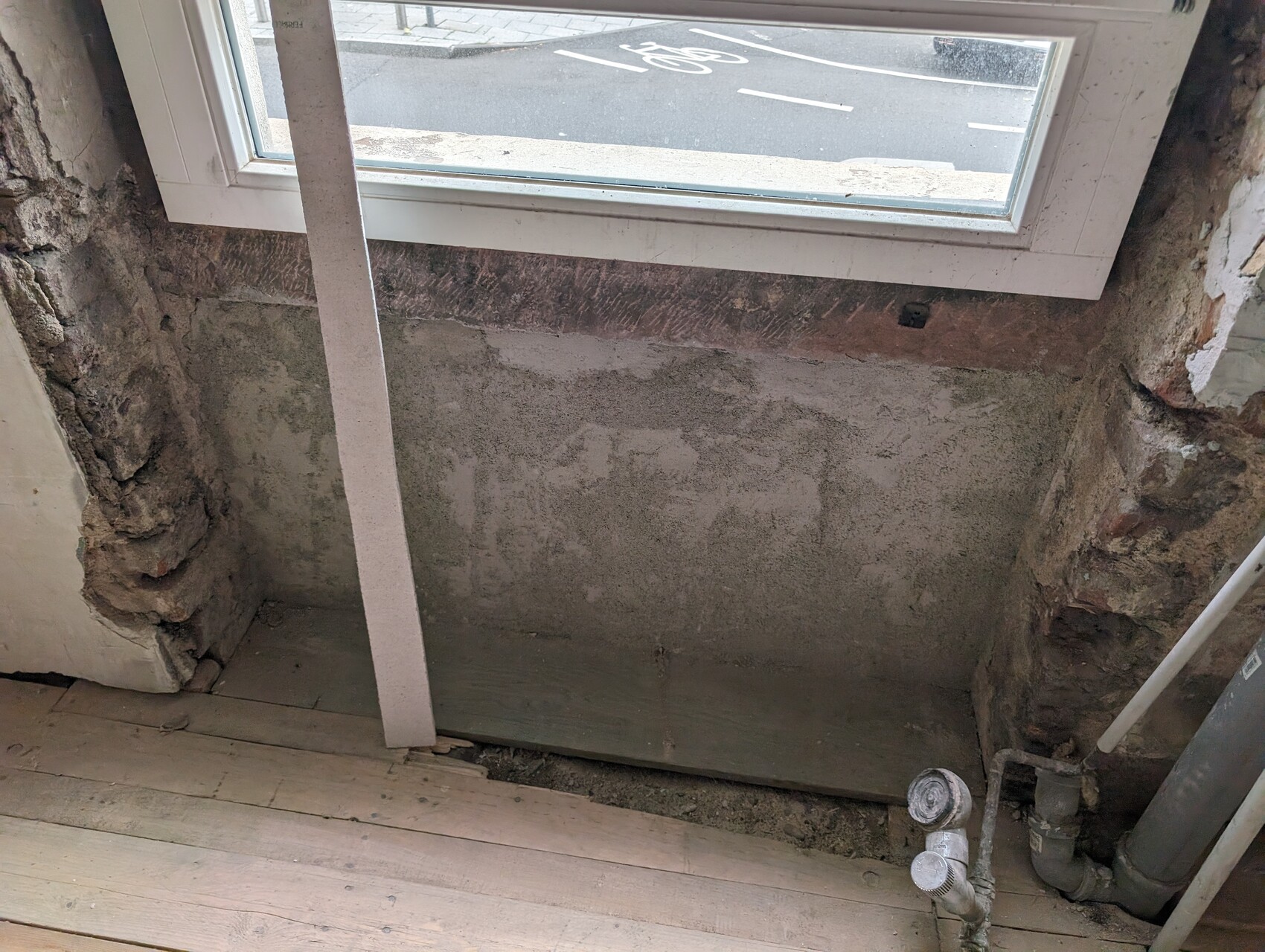 Renovierungsraum mit freigelegter Wand, Holzfußboden, Fensterfront und Rohren.