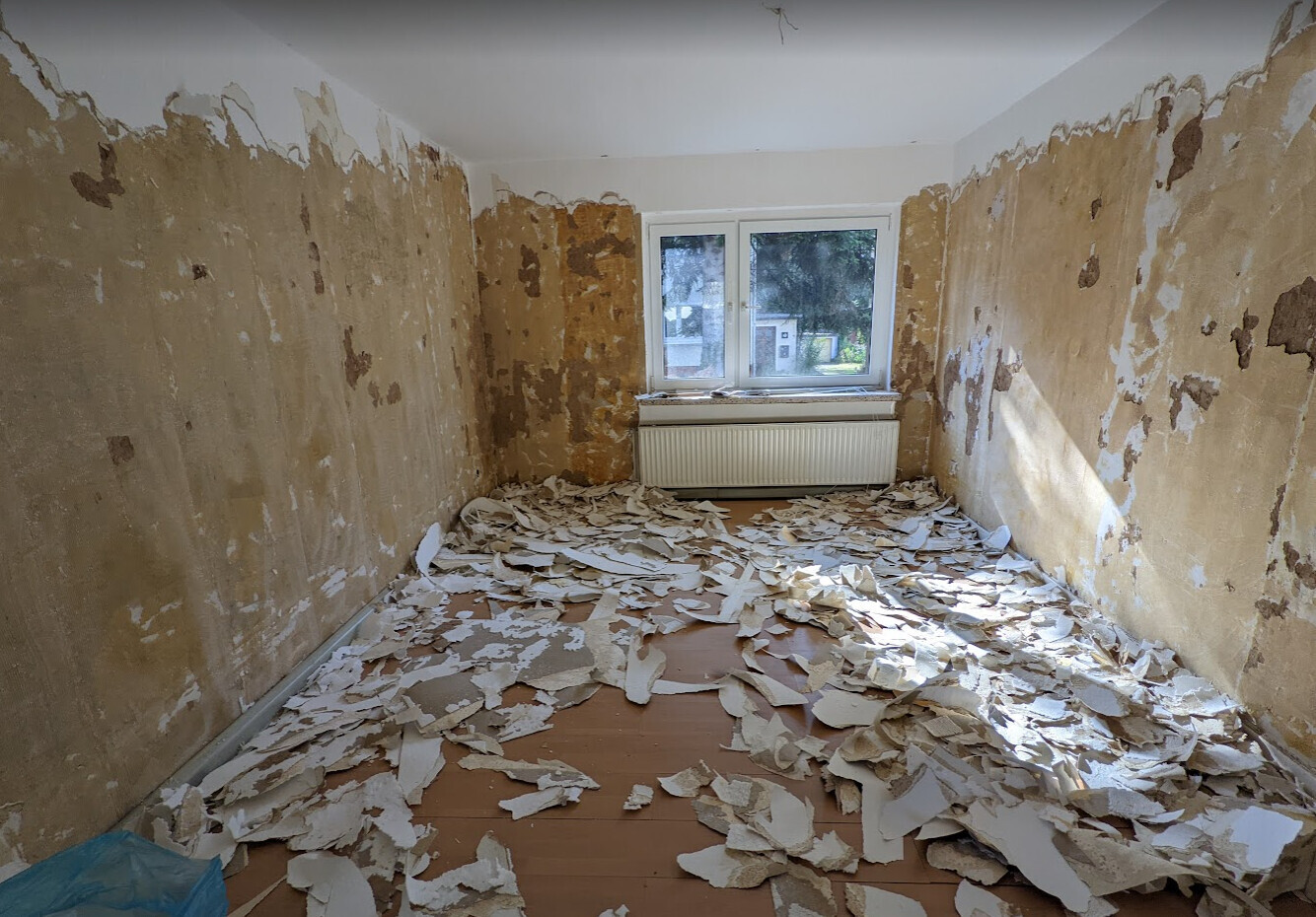 Raum mit abgeblätterter Wandtapete, Boden bedeckt mit Papierresten, Fenster mit Heizkörper darunter