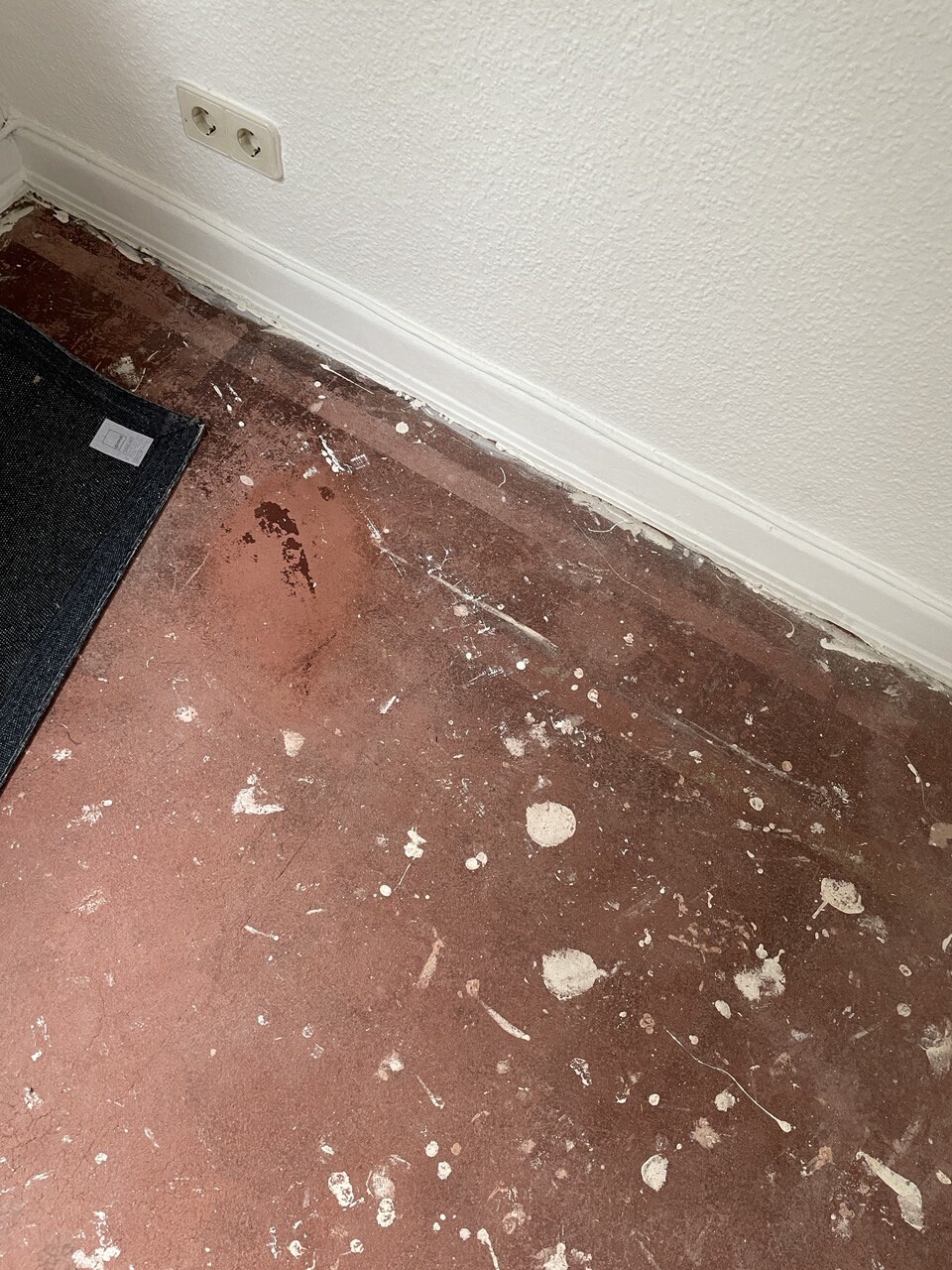 Eckansicht: roter Boden mit Farbflecken, weiße Wand, Sockelleiste und Doppelsteckdose