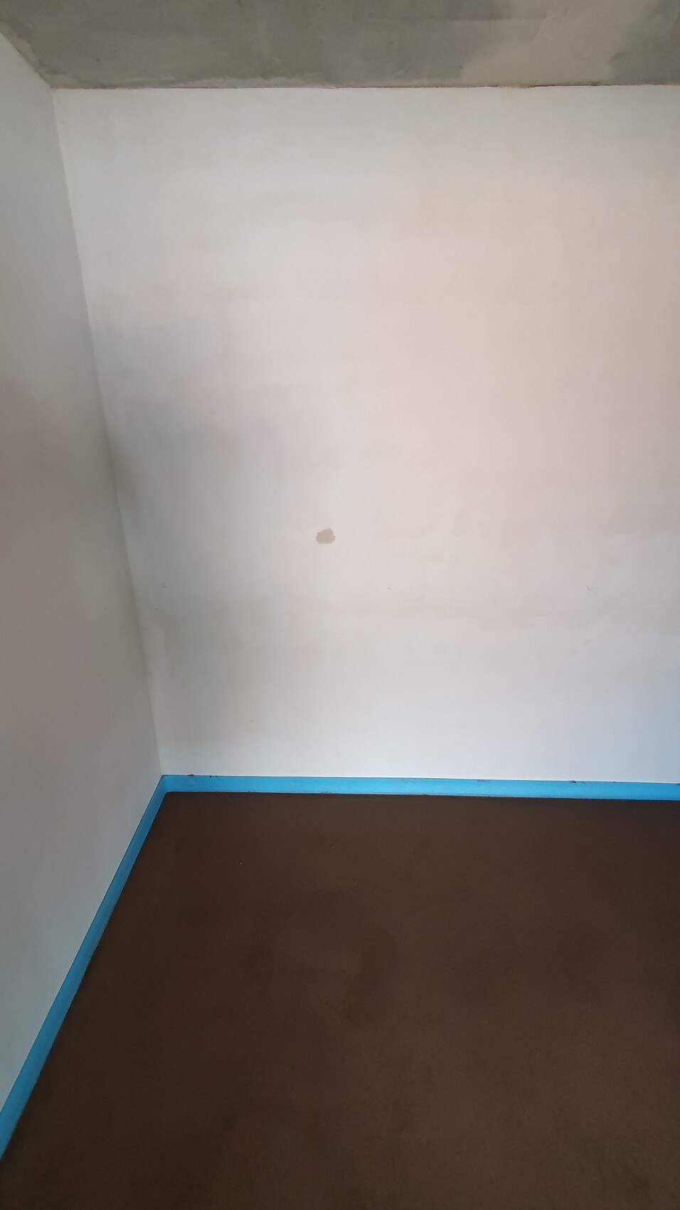 Ein Eckraum: weiße Wände, brauner Boden, blaue Sockelleiste; kleiner Fleck an der Wand.
