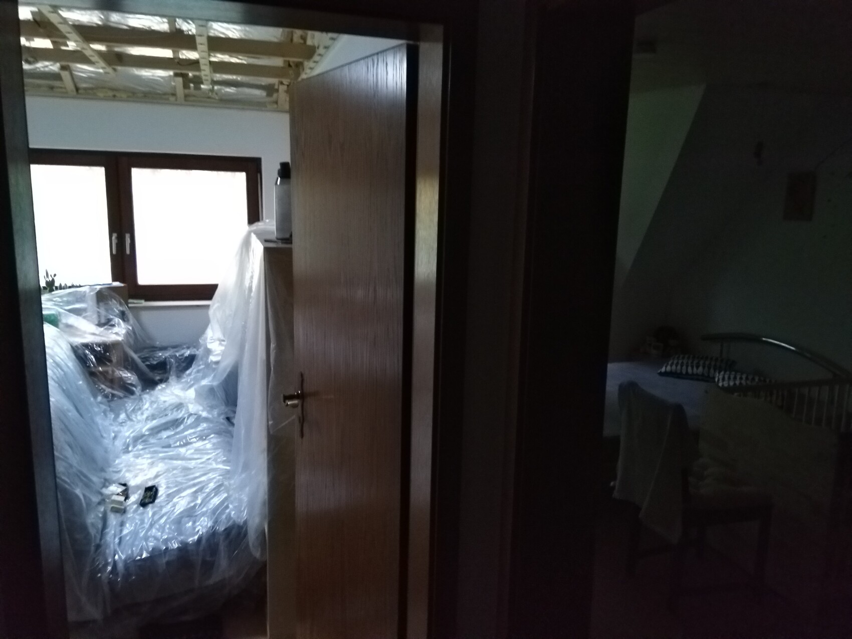 Dunkler Flur; links verpackte Möbel in Plastikfolie, rechts ein Bett im dunklen Zimmer.