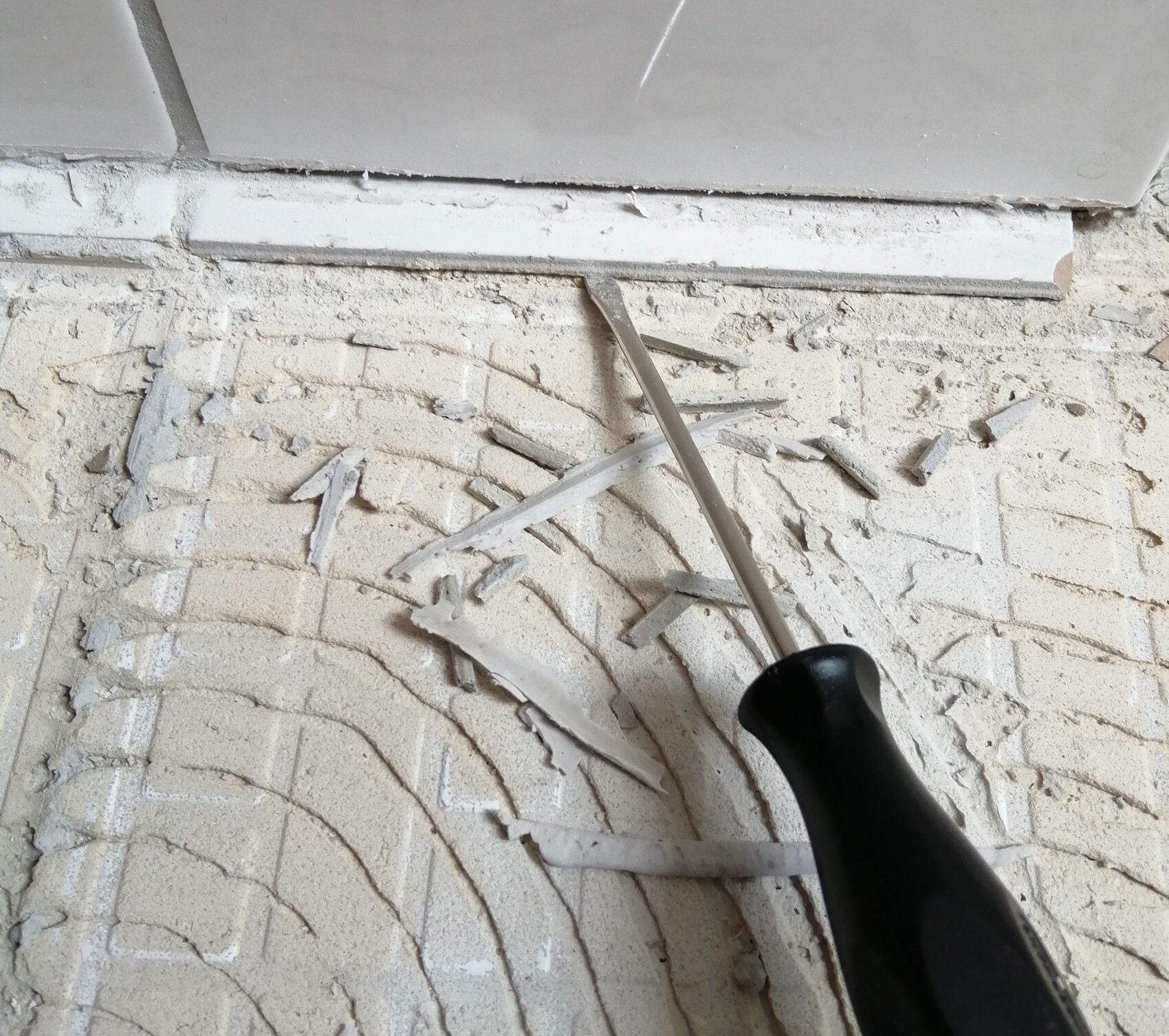 Schaber mit schwarzem Griff kratzt Verputz; Staub und Putzstücke liegen auf dem Boden.