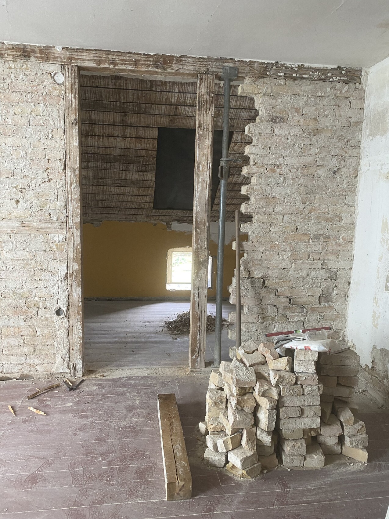 Baustellenraum mit Bruchsteinwänden, Ziegelstapeln, Holzbalken und Durchgang zu Raum mit gelber Wand